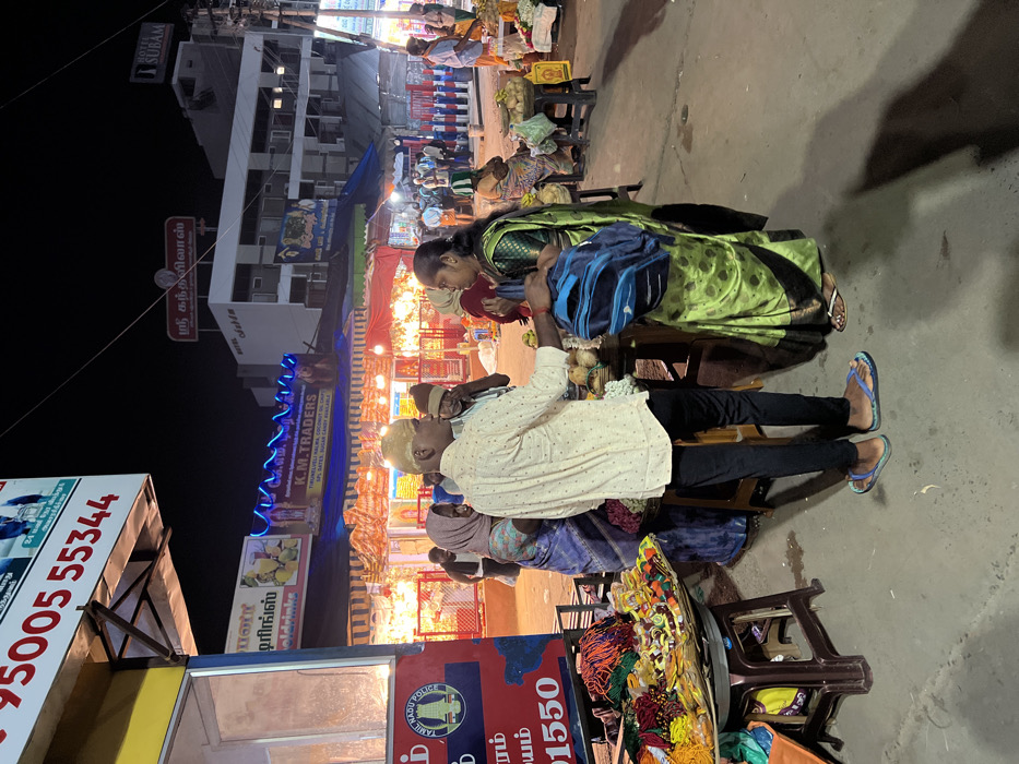 This is a nighttime scene depicting a bustling street or market in Palani, India. The image, originally rotated, shows a vibrant urban environment illuminated by artificial lights from shops and stalls.
In the midground, several individuals are gathered. A man wearing a light-colored, patterned shirt and dark trousers sits on a low stool, looking upwards and to the right, engaged in conversation. He wears blue flip-flops. Beside him, a woman in a green sari with gold patterns holds a blue backpack, looking downwards. Another person in a head covering and patterned attire is partially visible near them. Further back, more people are seen, some appearing to be vendors behind stalls laden with goods, including what look like fresh produce or coconuts.
The foreground features a street stall displaying various colorful items, possibly garlands or threads. Next to it, a sign with "TAMIL NADU POLICE" and the number "1550" is visible. Another sign displays a contact number: "95005 55344".
The background is dominated by multi-story buildings housing various businesses. Prominently featured is "HOTEL SUBAM" on a white building. Numerous signs in Tamil script and English indicate different shops. "K.M. TRADERS" advertises products like "TIRUNELVELI HALWA, COCONUT OIL CH," "SPL DATES, SUGAR CANDY BUNLABLE." Other signs include "ஸ்ரீ கந்தவிலாஸ்" (Sri Kanthavilas) and "டிரிங்ஸ்" (Drinks), along with "oldrinke" (likely cold drinks), suggesting beverage stalls. The street is decorated with string lights and blue neon lighting, contributing to the lively night market atmosphere. The overall scene captures a lively social interaction amidst street commerce.