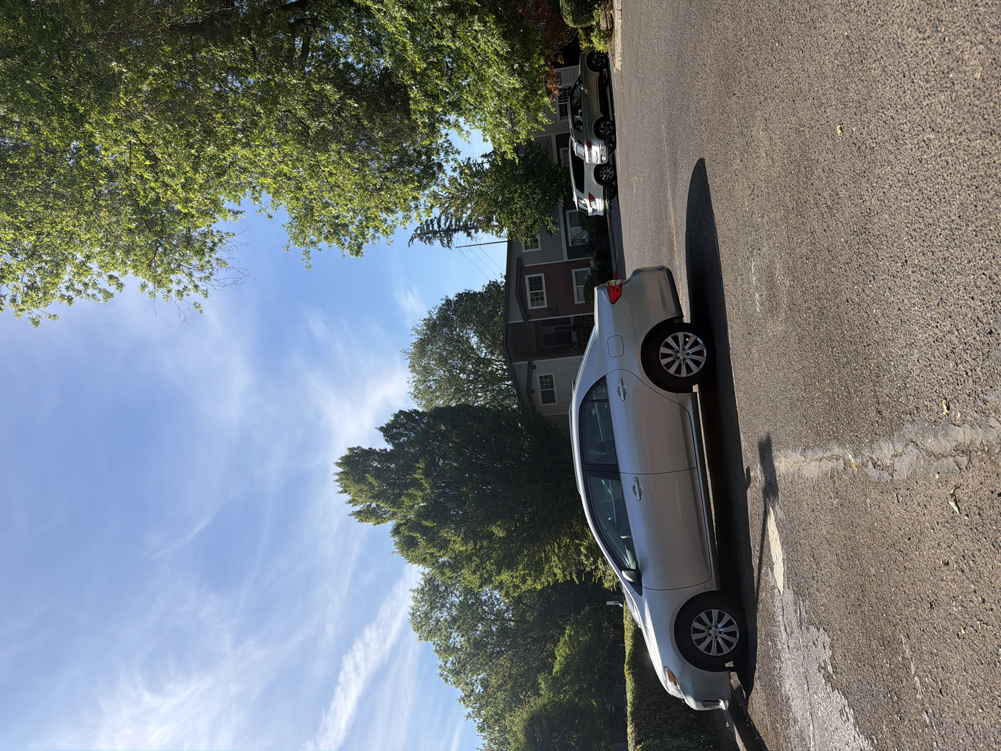 The image shows a street scene in Portland, Oregon. The perspective is tilted, suggesting the photo was taken from a low angle or the camera was rotated. In the foreground, a silver sedan is parked on the right side of the asphalt road. The car's tires and part of its body are visible. A long shadow from the car stretches across the road. Further down the street, two other cars are parked: a white SUV and a darker colored SUV. Behind the parked cars, houses with pitched roofs and windows are visible, interspersed with lush green trees. The sky is a bright blue with wispy white clouds, indicating it is daytime and likely fair weather. The presence of mature trees and the style of houses suggest a residential neighborhood. There is no visible text in the image.