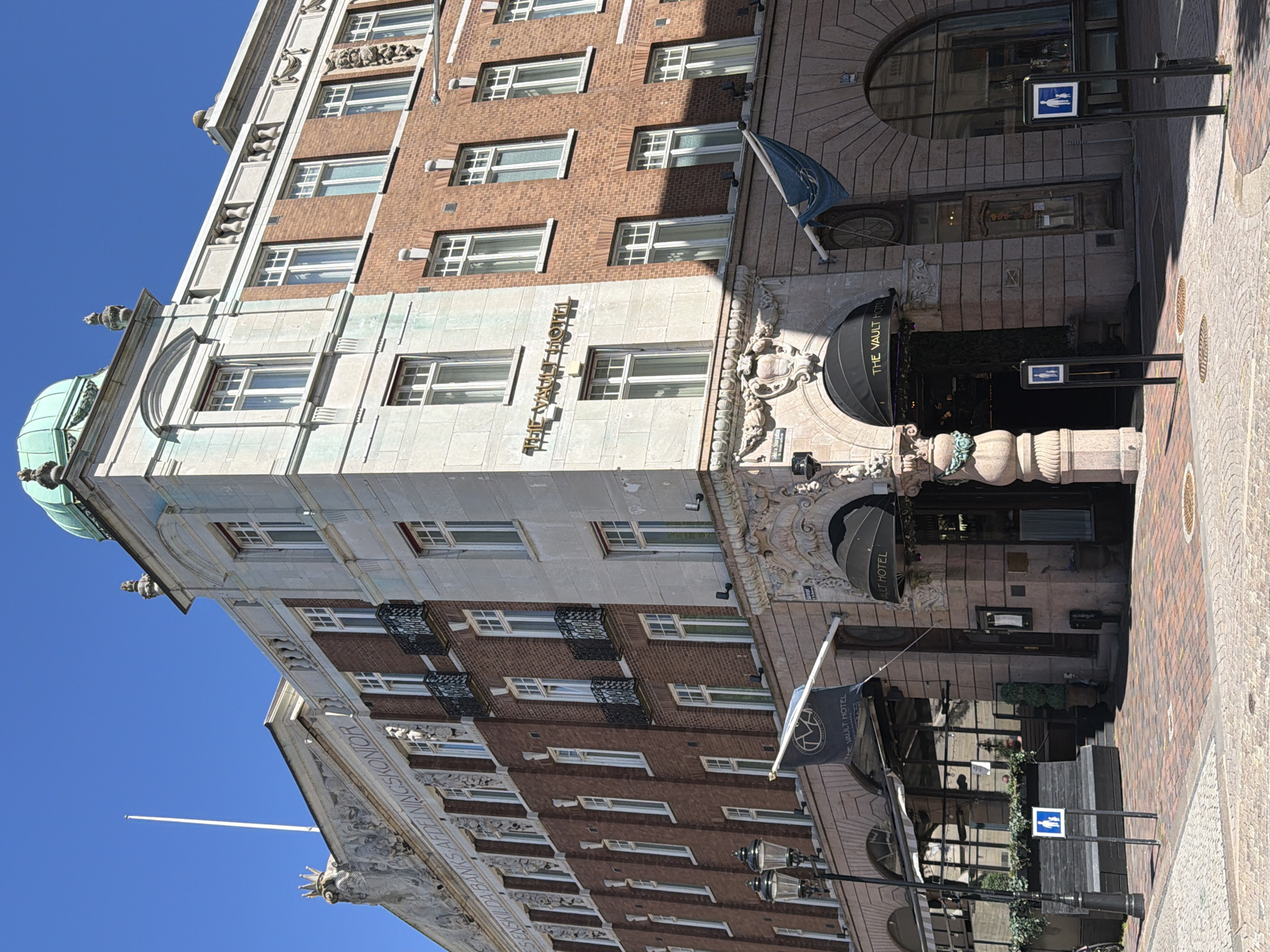 The image shows a facade of a building in Helsingborg, Sweden, with the inscription "ENSKILDA BANKS AFDILNINGSKONTOR" visible on the upper portion, indicating it was formerly a branch of a private bank. The building is primarily constructed of red brick and adorned with classical architectural elements, including ornate stonework, balconies with wrought-iron railings, and a green dome topping one section. Two flags are visible, one with the text "THE VAULT HOTEL" and another in blue. The ground level features entrances to "THE VAULT HOTEL", with awnings above them. Pedestrian crossing signs are positioned on the sidewalk. The sky is clear and blue, suggesting fair weather.