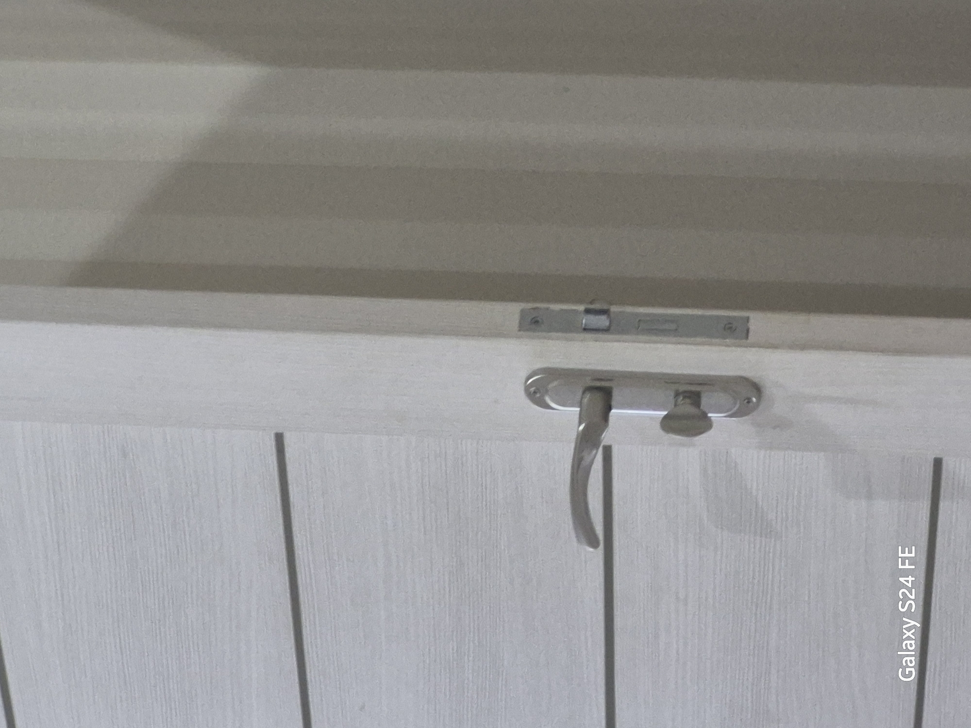 The image shows a close-up view of a light-colored wooden door with horizontal paneling. A silver door handle and a silver lock mechanism are visible on the door. The door is slightly ajar, revealing a vertical line of shadow on the adjacent wall, suggesting it opens inwards or outwards from the perspective of the camera. The lighting indicates an indoor environment, possibly a hallway or room. There is text in the lower left corner that reads "Galaxy S24 FE," which is likely a camera watermark. The image is devoid of people or any specific identifying landmarks that would place it in Chișinău, Moldova.