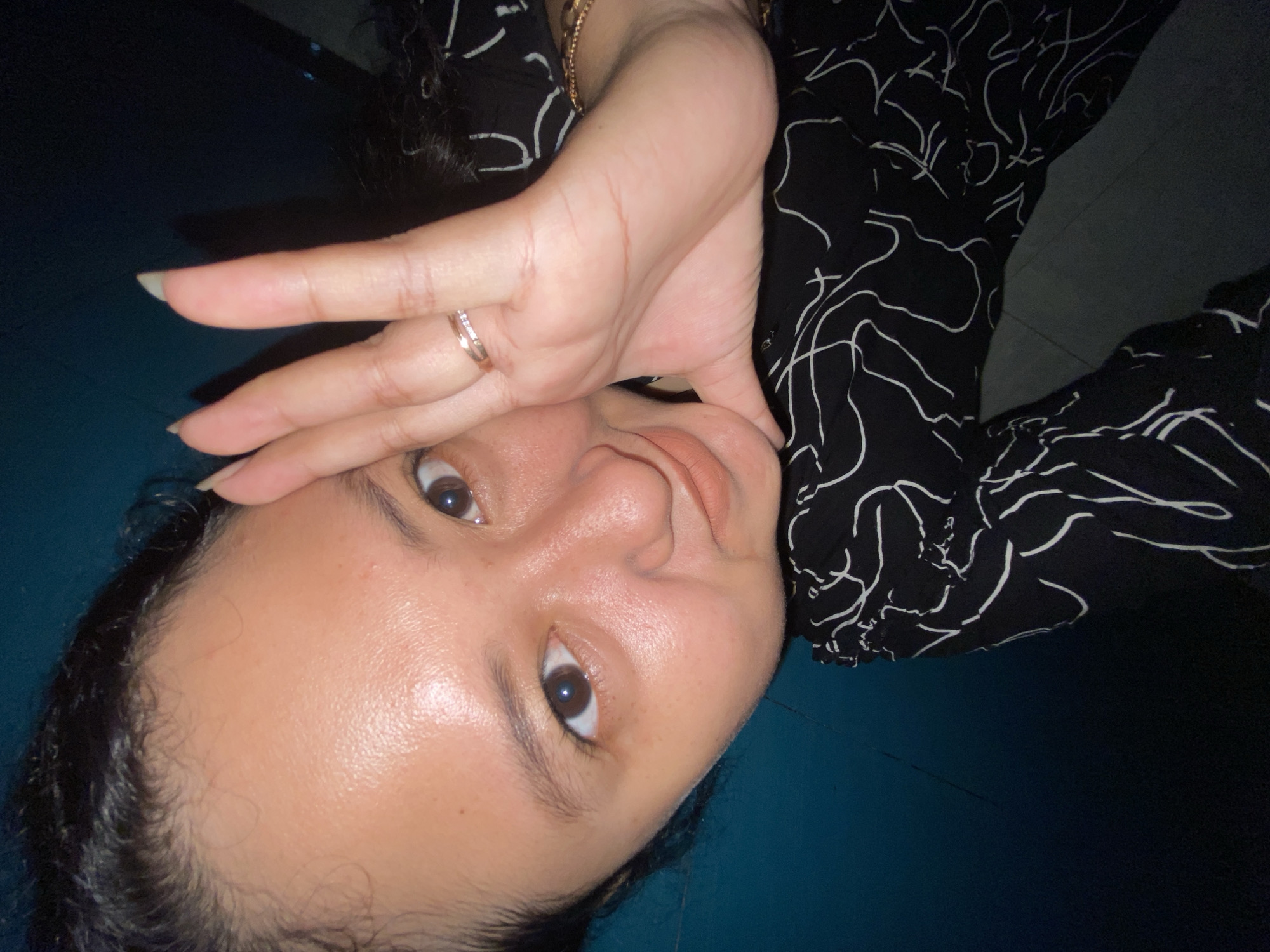 A close-up, selfie-style, portrait photograph shows a young woman from the chest up. Her dark hair is pulled back from her face, and she is leaning her right hand against her cheek. She is wearing a black top with a white, abstract pattern. She has a ring with small diamonds on her left hand. The background is a dark, teal-colored wall. The photograph appears to have been taken indoors in what might be an apartment in the city of Gelam, Indonesia.