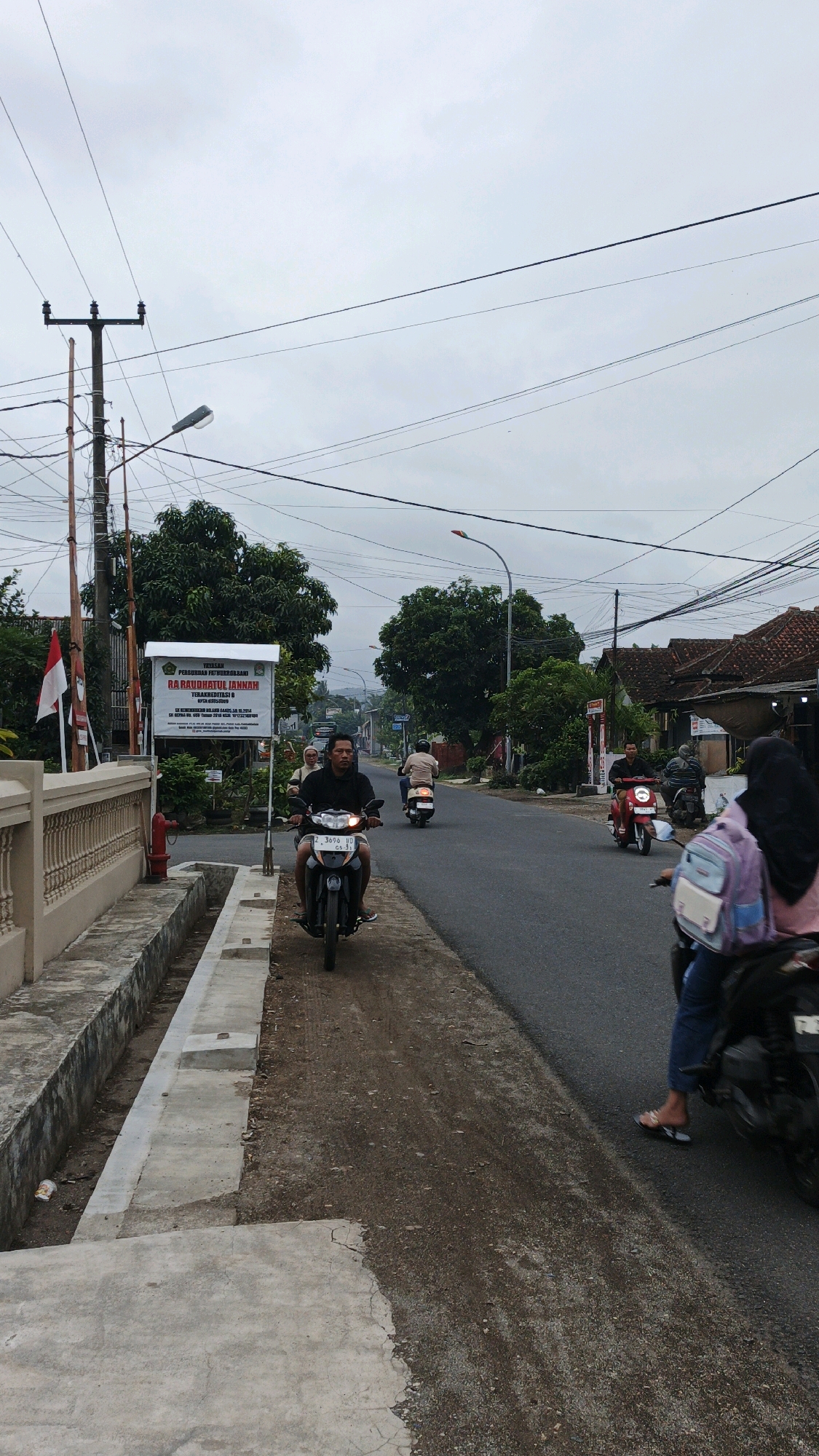The image depicts a street scene in Parigi, Indonesia, under an overcast sky. The foreground features a concrete sidewalk with a drainage ditch running alongside it. A man is riding a scooter towards the viewer. He is wearing a black hoodie and dark pants. His scooter has a license plate that reads "3096 UD GS.3.".
Further down the road, other individuals are seen riding motorcycles. One person, wearing a light-colored shirt, is riding a scooter away from the viewer. Another scooter, colored red, is also moving away from the viewer. On the right side of the road, a person wearing a hijab and jeans is riding a scooter, and a second person is seated behind them. The person in the back has a pink and blue backpack.
On the left side of the road, there is a white sign with text in Indonesian, and a red and white flag is attached to a pole nearby. The background shows a line of trees and buildings with red-tiled roofs. Power lines crisscross the sky, supported by utility poles. The overall environment appears to be a residential or semi-urban area.