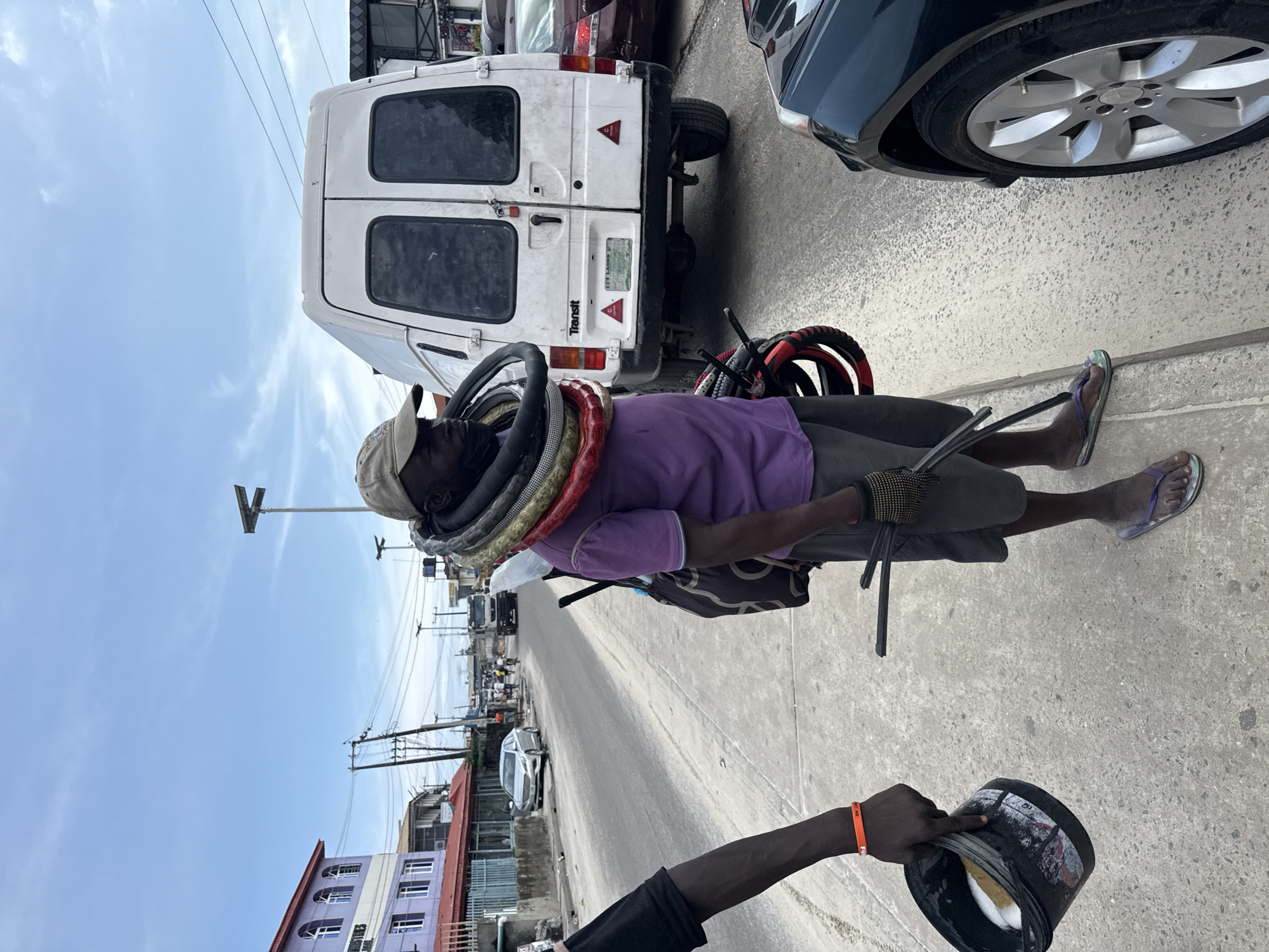 The image depicts a street scene in Epe, Nigeria, under a cloudy sky, suggesting it is daytime. The foreground is dominated by a person wearing a purple shirt, grey shorts, a beige baseball cap, and flip-flops. This individual is carrying a stack of what appear to be inner tubes or rubber rings around their neck. They are also wearing a glove on their right hand and holding some metal rods.
To the right of the person, another arm is visible, holding a black bucket. The bucket has some text and images on its side, and it appears to contain some white material. An orange bracelet is worn on the wrist of this second individual.
In the background, several vehicles are parked and driving on a paved road. Notably, a white van with "Transit" written on its back is prominent. Other cars, including a dark-colored sedan with silver rims, are also present. Buildings line the street, with one appearing to be a multi-story structure with purple and white facades and arched windows. Power lines are visible across the sky, a common sight in many urban and semi-urban areas.
The overall impression is a snapshot of daily life on a Nigerian street, possibly capturing a vendor or someone offering services related to the items they are carrying, or perhaps a scene of informal commerce. The presence of the bucket suggests a collection or donation element. The lighting and sky indicate fair weather during daylight hours.