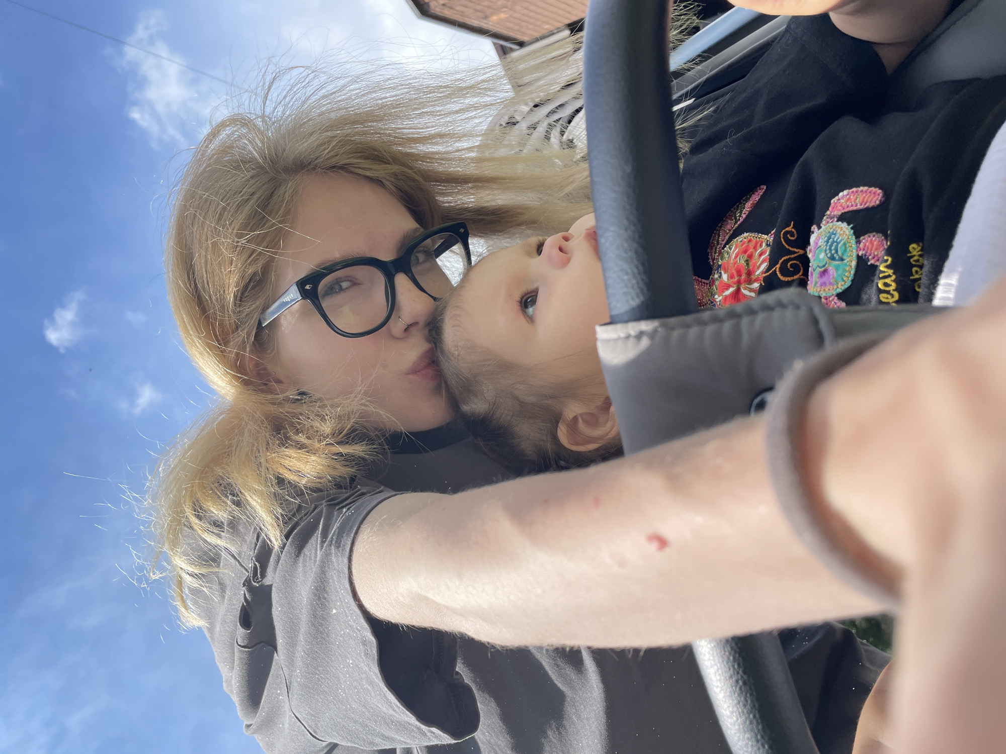 A blonde-haired person wearing glasses leans over a baby in a stroller, kissing the baby's forehead. The background shows a bright blue sky with scattered clouds and a brick building. The person is wearing a dark t-shirt with subtle glittery details. The baby is wearing a black t-shirt with colorful embroidered sea creatures. The scene appears to be outdoors, possibly on a sunny day. The image does not contain specific landmarks that would allow for definitive identification of the city of Izhevsk, Russia.