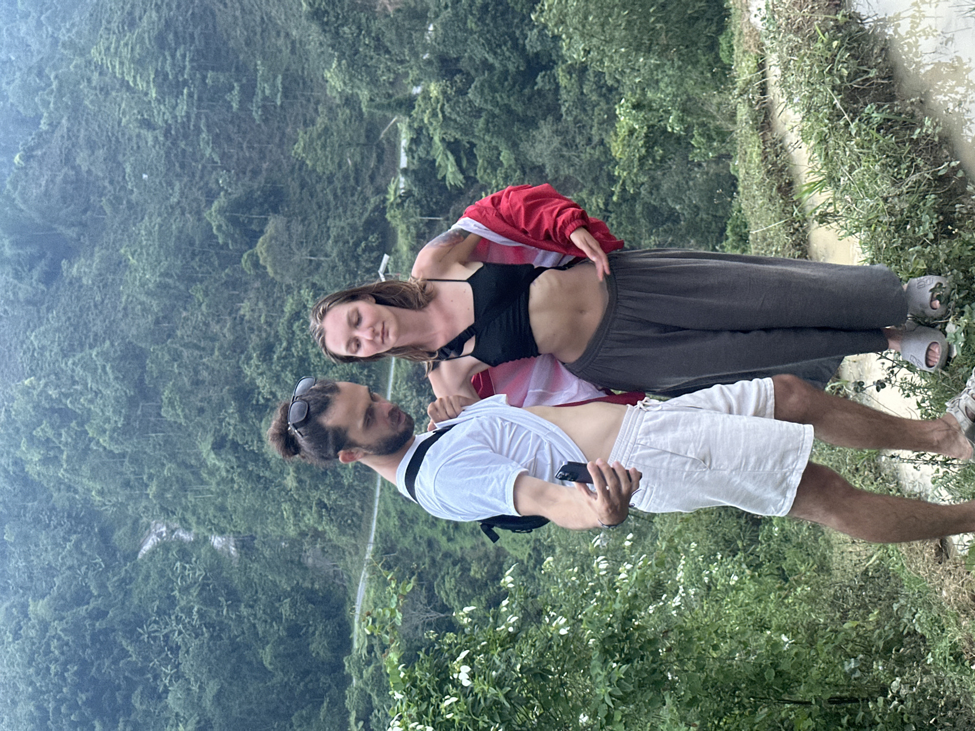 The image, when correctly oriented, depicts two individuals standing outdoors against a backdrop of a vast, lush green landscape in Nậm Dịch Commune, Vietnam.
On the left, a woman with light brown hair looks towards the man. She is wearing a black crop top, dark grey wide-legged pants, and grey slides. A red jacket is draped over her shoulders, revealing a white garment beneath. Her expression appears calm or contemplative.
On the right, a man with dark hair tied in a bun and sunglasses resting on his head looks at the woman. He has a beard and is dressed in a white t-shirt and white shorts. He is holding a black smartphone in his right hand and has a black crossbody bag across his chest. His gaze is directed towards the woman.
The setting is characterized by dense, verdant forest or mountainous jungle that recedes into a hazy distance, suggesting a cloudy or misty day. A faint outline of a road or path can be seen winding through the distant hills. In the immediate foreground, to the right of the people, there is earthy ground covered with small green plants, and a small body of water or a puddle reflecting light. The scene is captured from a high vantage point, offering an expansive view of the natural terrain. The time of day appears to be daytime, with diffused light indicating overcast weather. There is no visible text in the image.