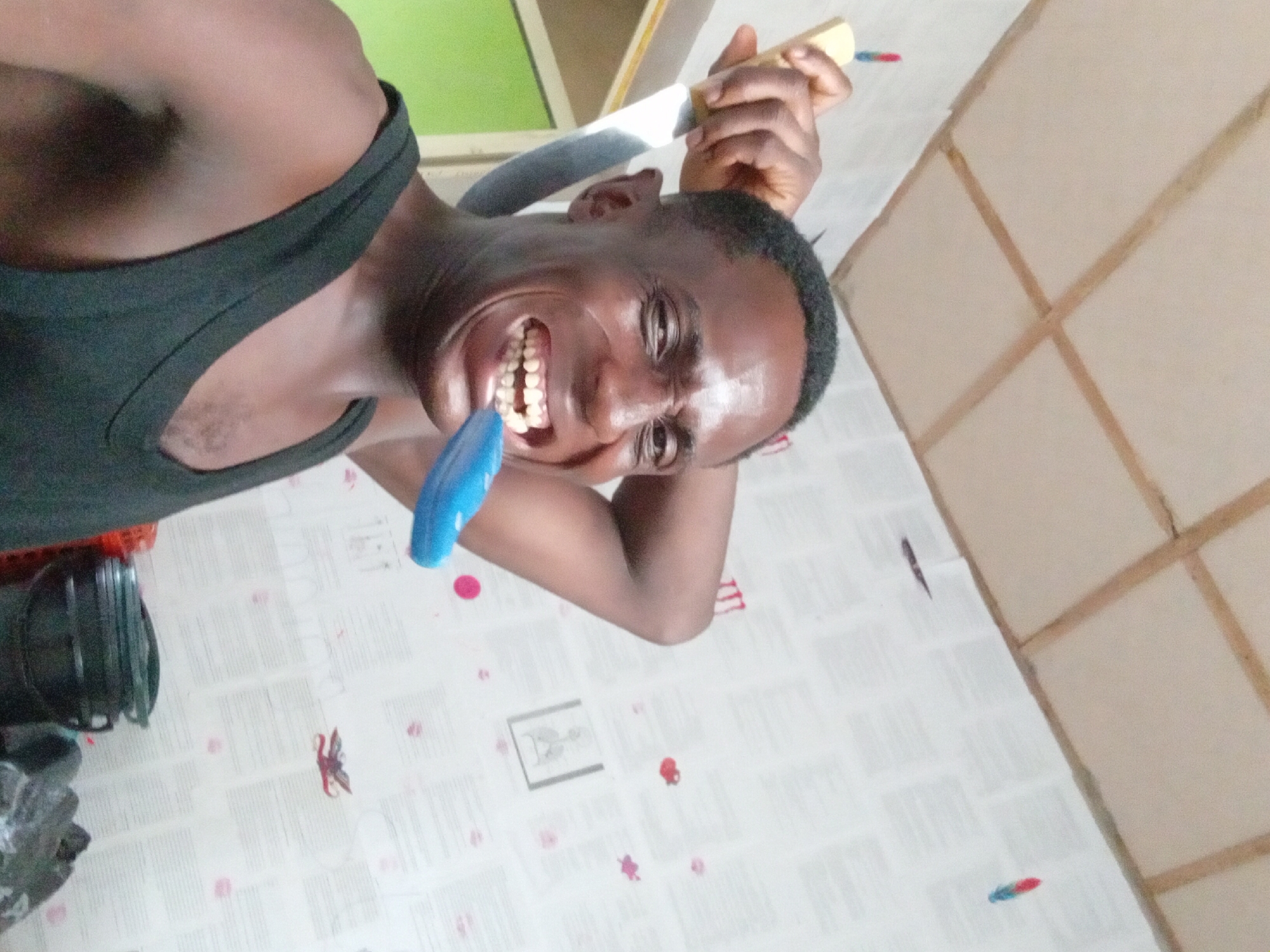 A man stands indoors, wearing a black tank top. He holds a large kitchen knife above his head with his right hand, while a blue object is held in his mouth. The man is facing the camera and grinning widely, displaying his teeth. The background shows a wallpaper with various objects printed on it, and there's a wall with a grid-like pattern on the right side of the frame. A green surface can be seen through a window in the background. A black object sits on the left side of the scene. The location is Akwete, Nigeria.