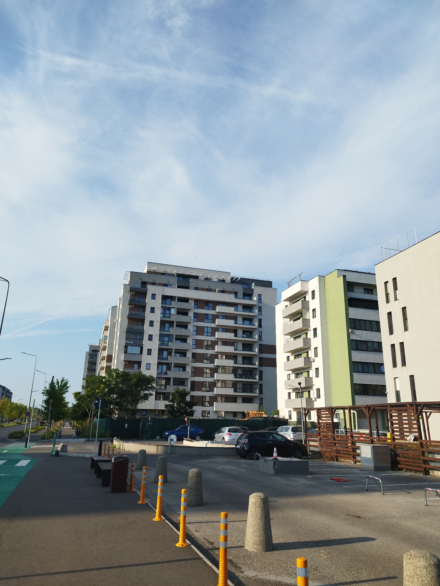 The image shows a modern residential area in Bucharest, Romania, featuring several multi-story apartment buildings. The buildings are contemporary in design, with balconies and large windows. In the foreground, there is a paved street with yellow and white traffic cones, bollards, and benches. A green pedestrian and bicycle lane is visible on the left side of the street. Several cars are parked in a lot to the right, adjacent to a wooden fence. The sky is partly cloudy with a light blue hue. The scene depicts a typical urban environment with residential structures and public amenities.
