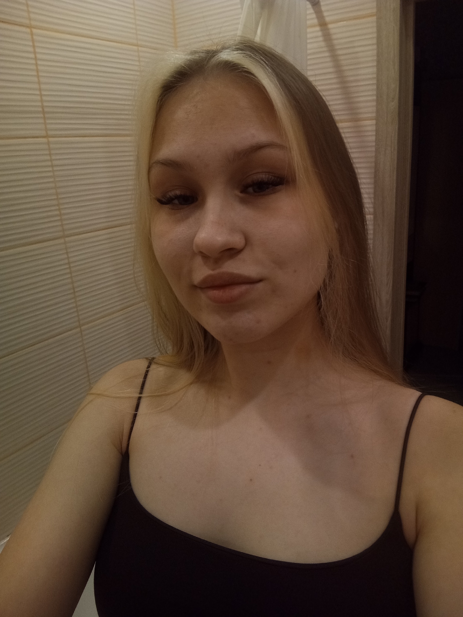 A young woman with blonde hair and brown eyes is facing the camera, wearing a dark-colored tank top. She appears to be indoors, standing in a bathroom. The background shows beige horizontal tiles on the wall and a partly visible wooden door. The photo appears to have been taken in a dimly lit environment. There is no evidence in the image to indicate that it was taken in Krasnodar, Russia.