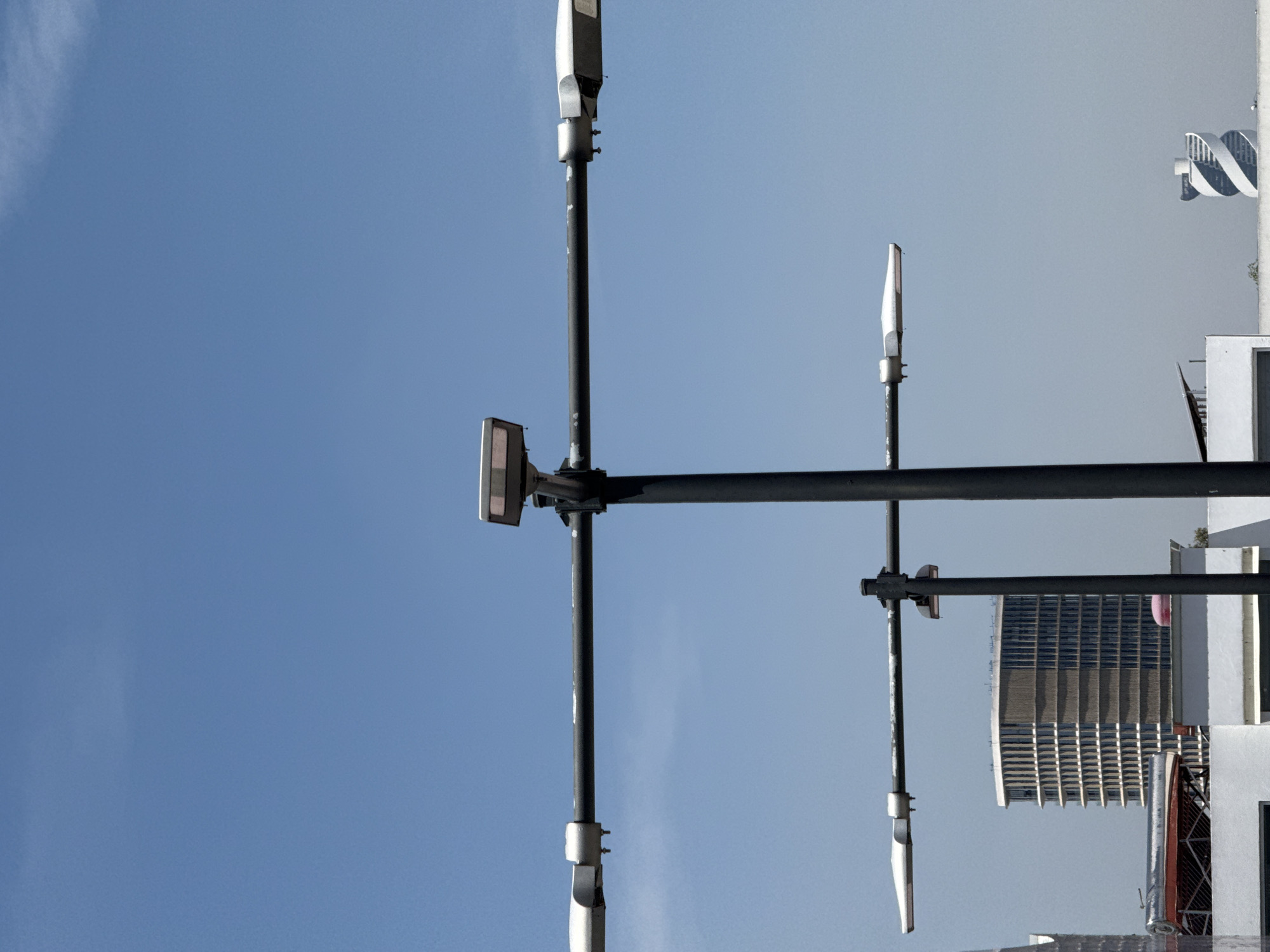 The image captures an upward-looking view of urban infrastructure and architecture against a clear blue sky. Dominating the foreground is a tall, dark-colored utility pole, likely a street light or a similar support structure. This central pole features two prominent horizontal crossbars, from which multiple modern light fixtures extend. These fixtures are sleek, elongated, and white or light gray, suggesting contemporary design, possibly LED streetlights. Another shorter, similar pole structure is partially visible on the right side. The poles show some signs of wear, with slight discoloration or chipped paint.
In the background, under a bright, cloudless sky, several buildings are visible. To the upper right, a distinctive white architectural element with a spiral or twisted design stands out, resembling a modern sculpture or part of a unique building façade. Below this, a large, multi-story building with a uniform grid of dark windows and strong vertical lines across its facade is prominently featured. Additional white or light-colored building structures are visible along the right and bottom edges of the frame.
The time of day appears to be daytime, likely morning or afternoon, due to the bright, clear blue sky and the even illumination without harsh shadows. The weather is fair and sunny, with only a few faint wisps of clouds barely perceptible. No specific activity or event is taking place, as the image is a static capture of the urban environment. There is no visible text. The modern architecture and infrastructure align with the provided context of Puebla, Mexico.