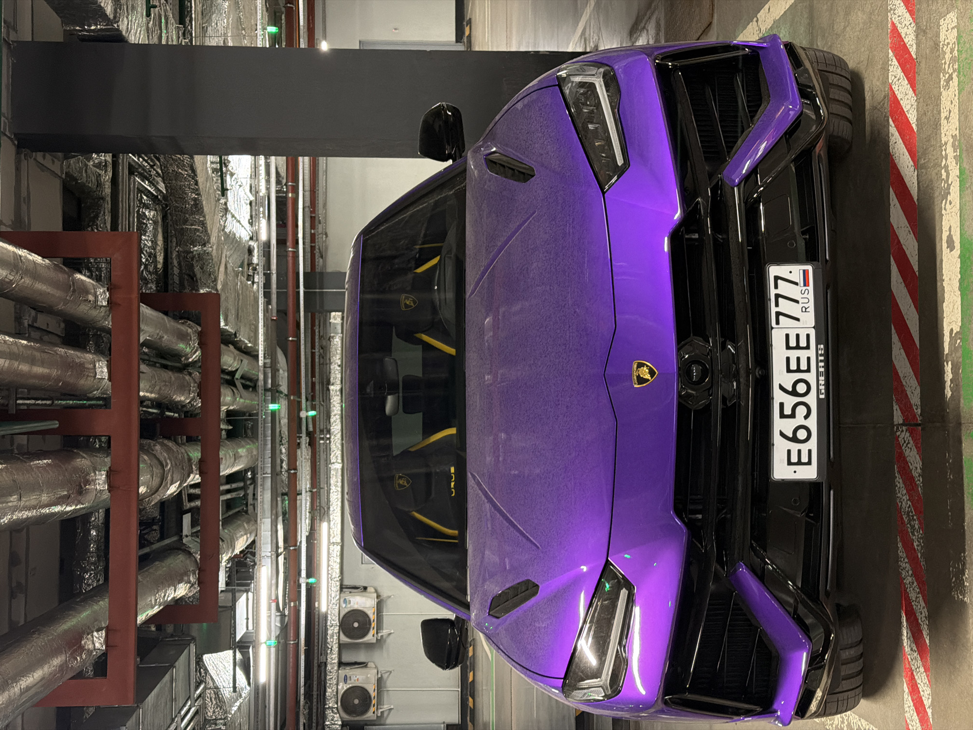 The image depicts a purple Lamborghini Urus parked in what appears to be an underground parking garage. The car has a Russian license plate. The environment includes overhead pipes and ventilation systems, indicative of a utilitarian space. The location is indicated by the Russian license plate, suggesting the city of Moscow, Russia.