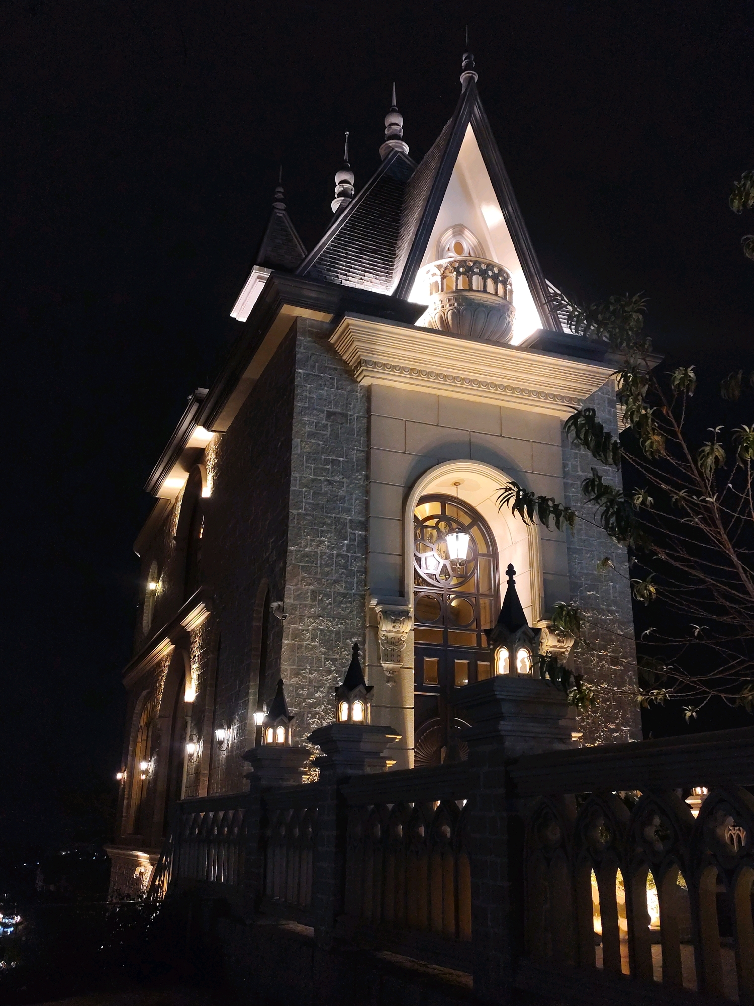 A tall, gothic-style building is illuminated at night in Da Lat, Vietnam. The building features pointed spires, arched windows, and stone masonry. It is set against a dark sky, with a few trees visible on the right side of the frame. The building is lit with warm, yellow lights, highlighting its architectural details. A decorative stone fence or railing is in the foreground, also illuminated from behind by the building's lights. The scene suggests a historical or fantastical setting, characteristic of some tourist attractions in Da Lat.