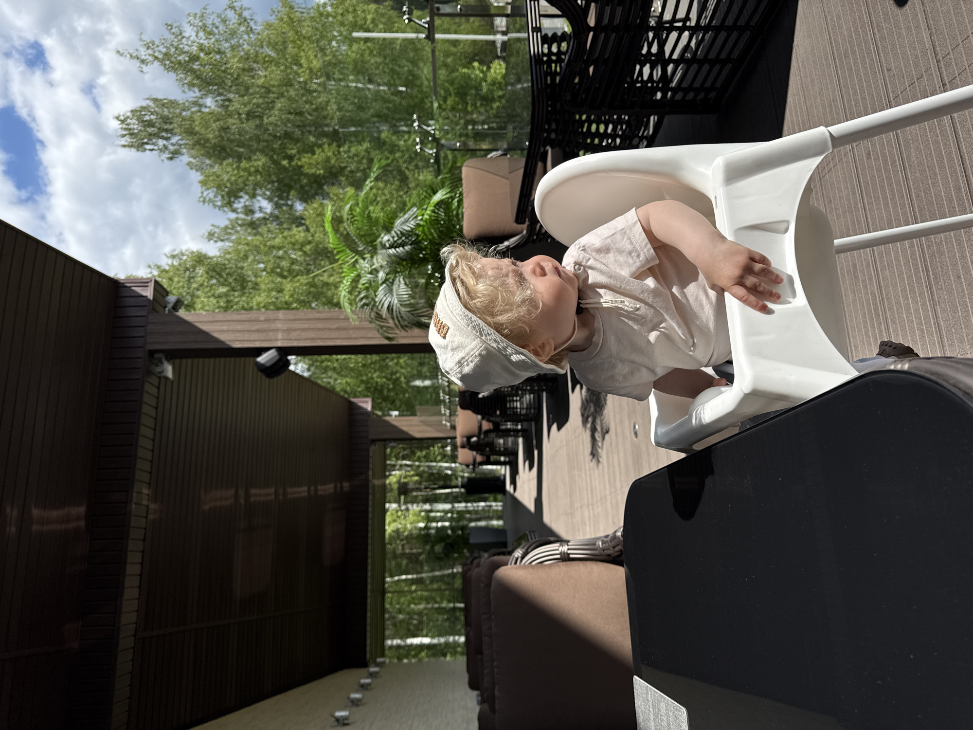 An outdoor scene in Dobrograd, Russia, captures a young child seated in a white high chair on what appears to be a sunny patio or terrace. The child, with light blonde curly hair, wears a light-colored t-shirt and a matching cap, looking upwards and slightly to the left with a calm or curious expression. Their right arm rests on the side of the high chair.
The setting includes a modern building with dark, vertically paneled siding on the left, receding into the background. Lush green trees are visible against a bright blue sky dotted with white clouds, suggesting a pleasant, clear day. In the middle ground, there's outdoor patio furniture with dark frames and brown cushions, indicating a comfortable lounge or dining area. A paved ground with small embedded lights runs along the base of the building, adding to the refined outdoor ambiance. A dark, reflective surface, possibly a table or an appliance, is prominent in the foreground. No discernible text is visible in the image.