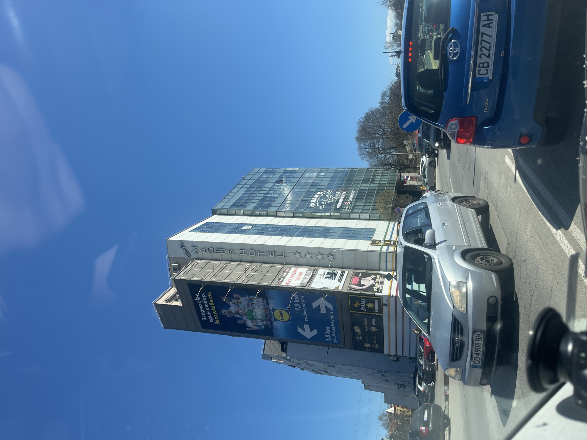 The image shows a street scene in Sofia, Bulgaria, featuring a car in the foreground with license plate "CO 4908 BH", and a blue Toyota behind with license plate "CB 2277 AH". To the left, there is a multi-story building that appears to be the "Magus Hotel" with an adjacent sign promoting rental spaces. There is a billboard pointing towards Lidl, Palms, and Merkur, with directions to each of the destinations. Above these signs is a large glass office building, with the signage of "ФЛАЙС" and “SPORTDEPOT” on the building, in addition to various other businesses. The sky is clear and blue. Traffic appears to be stopped.