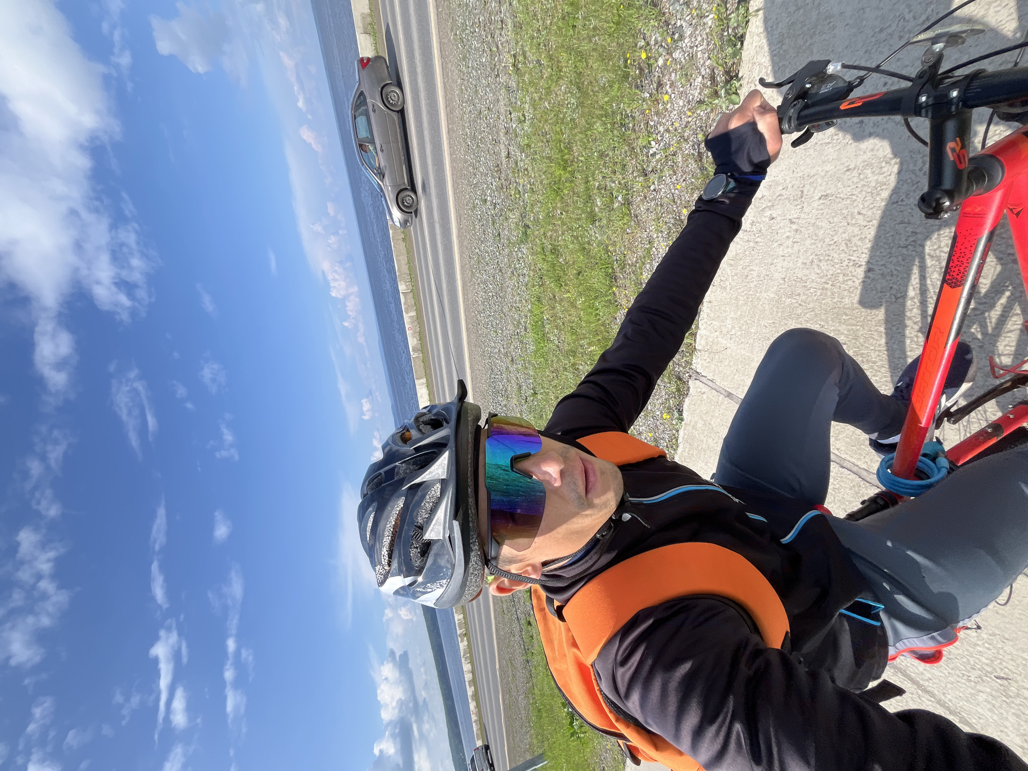 A man is riding a red bicycle on a paved path next to a road. He is wearing a black long-sleeved shirt, grey pants, a black helmet, and mirrored cycling glasses. An orange backpack is on his back. The road has a gray car driving on it. Beyond the road is a body of water with a cloudy blue sky above. The location is likely the embankment of the Kama River in Chaikovsky, Russia, as the scene depicts a body of water, a road, and a paved path suitable for cycling, consistent with the known geography of the city. The man is looking forward, appearing to be in motion.