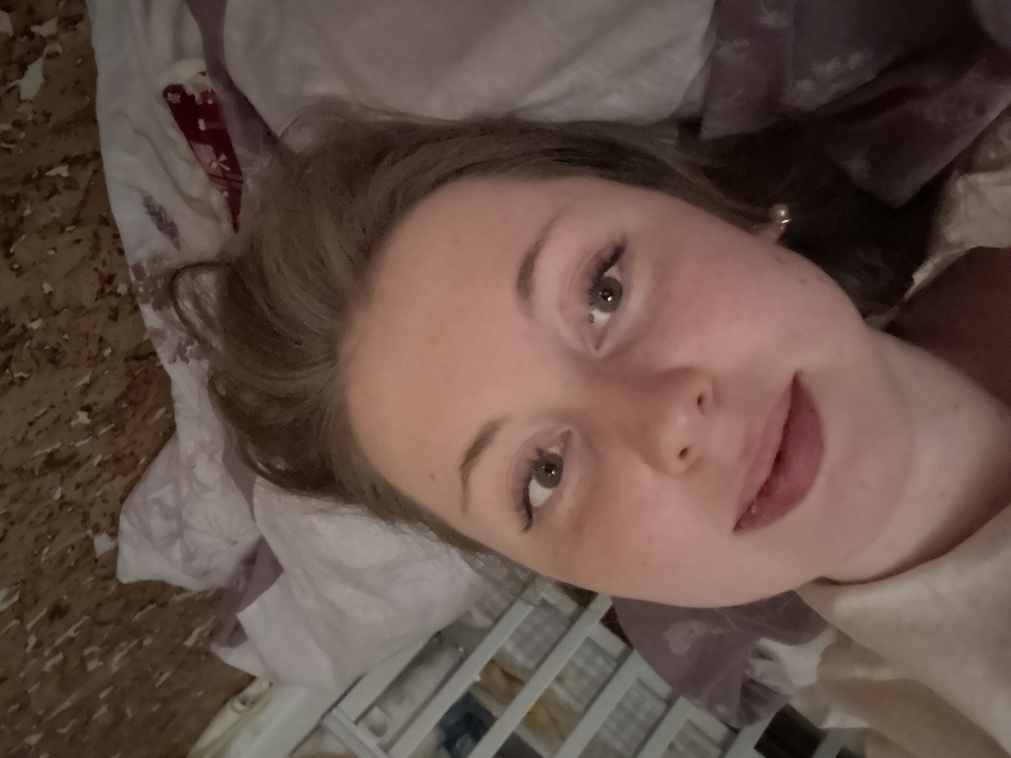 A young woman is captured in a close-up, slightly overhead selfie, lying down in what appears to be a bed. She has light skin, light brown hair, and direct eye contact with the camera, accompanied by a subtle, relaxed smile. Her eyes are a light brown or hazel, enhanced with mascara, and her lips have a hint of color. A small, light-colored stud earring is visible in her right ear. She is resting on white bedding, which has faint patterns and includes a section of red patterned fabric folded within. To her left, a textured, mottled light brown surface, possibly flooring, is visible. A grid-like storage unit or shelving is partially visible in the bottom right corner of the frame. The lighting is soft, suggesting an indoor setting. No text is visible.
