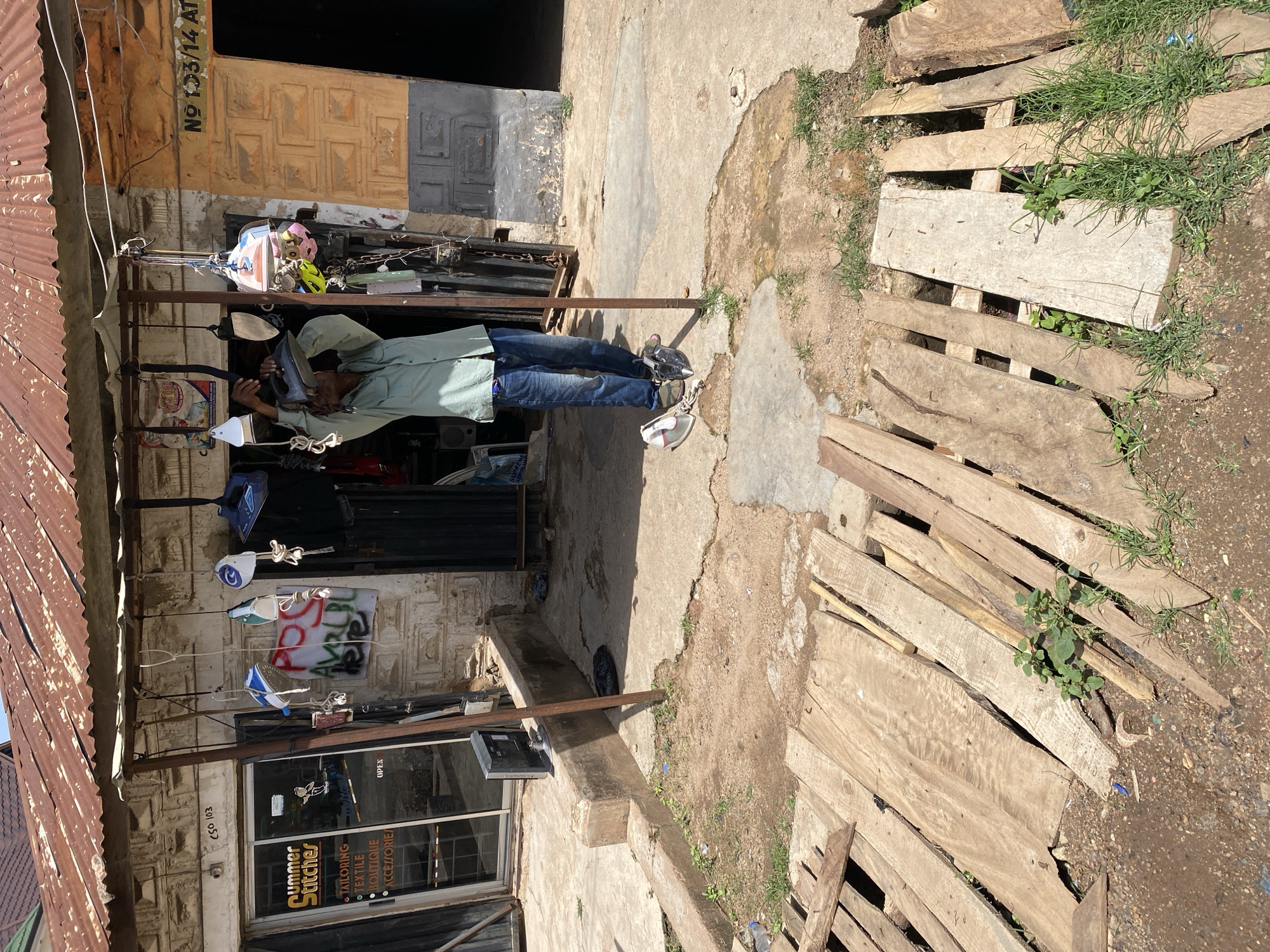 The image shows a street-level view of a small shop in Jos, Nigeria. The shop has a corrugated iron roof and a brick facade. Several items, including clothing and what appear to be irons, are hanging outside the shop. A man is standing in front of the shop, appearing to be handling an iron. The ground in front of the shop is a mix of concrete and dirt, with some wooden planks laid out. The shop's signage, "Summer Stitches," suggests it is a tailor or clothing store. The overall impression is one of a busy, commercial area in a Nigerian city.