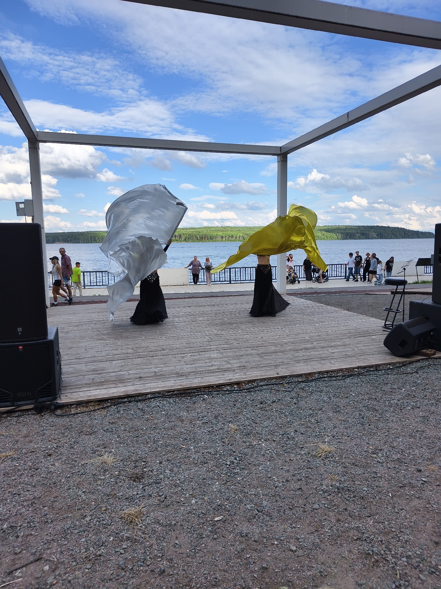In the city of Unknown, Unknown, an outdoor performance stage is set up on a wooden deck. Two performers in black dresses are dancing, one holding a large silver fabric and the other a large yellow fabric, both creating dynamic visual effects. Behind them, a body of water stretches out, with a treeline in the distance. A few spectators are visible along the waterfront, and some large speaker boxes are positioned on either side of the stage. The sky is blue with scattered white clouds.