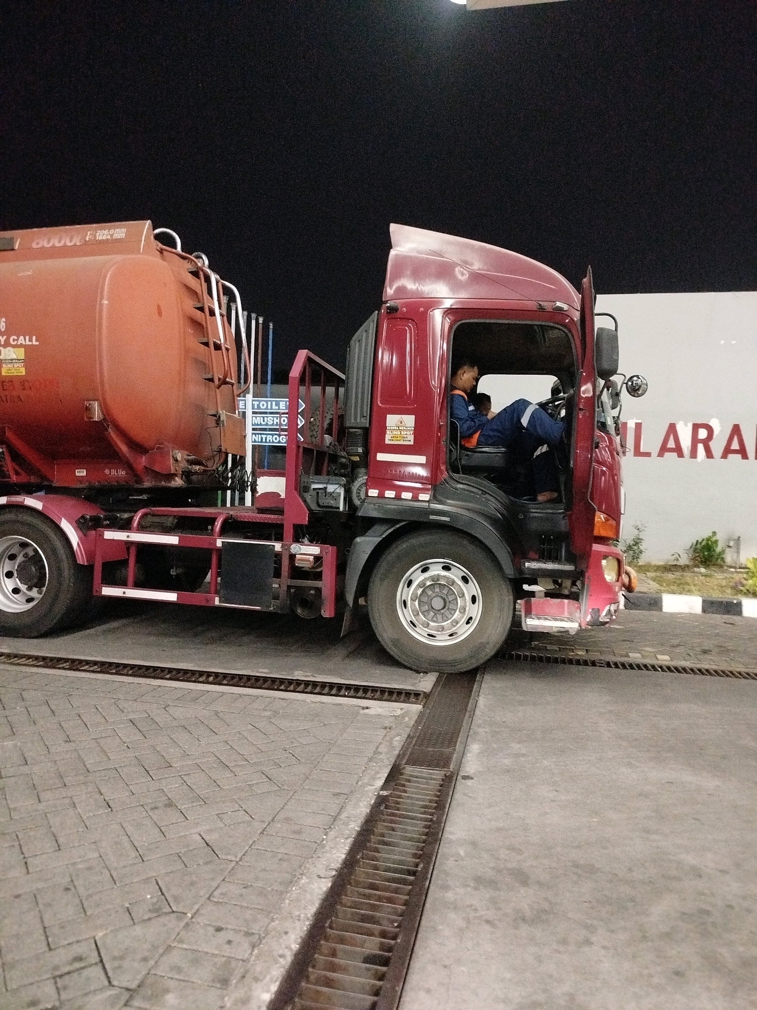A stationary, large, red fuel tanker truck is parked at a gas station in Surabaya, Indonesia. The truck's engine is running, and the driver, a male wearing a blue uniform with reflective stripes, is seated in the cab with the door open. He appears to be looking at his phone. Another person, also in a blue uniform, is partially visible behind the driver. The gas station has a paved surface with drainage grates and a white wall in the background with red lettering that reads "LARANG" (Indonesian for "forbidden" or "prohibited"). The time of day is likely night, as indicated by the dark sky and artificial lighting. The tanker itself is orange and shows some wear and tear.