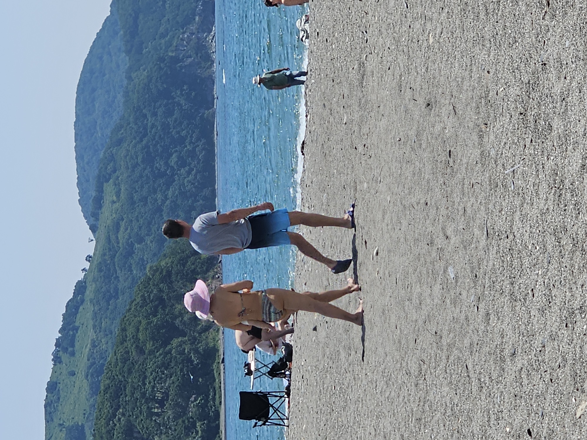 A bright, sunny day at Nakhodka, Russia's Primorsky Krai, as several people are on a pebble beach with the Sea of Japan in the background. The sea is a vibrant blue, with light waves breaking near the shore. Lush green, tree-covered hills rise up from the coastline, forming a dramatic backdrop to the scene.
In the foreground, a couple walks away from the camera. The woman wears a pink sun hat and a striped bikini. The man, to her right, is in blue swimming trunks and a grey t-shirt. Their presence suggests a leisurely day by the water.
Further back, near the water's edge, a man in a straw hat and green t-shirt stands facing the sea. To his right, a person is partially visible, sitting on the beach. In the mid-ground, a black camping chair and a small folding table are set up on the sand, suggesting a spot for relaxation or a picnic. The presence of other individuals and beach paraphernalia indicates a public area used for recreation.
The beach itself is composed of small pebbles and sand, with some scattered debris. The overall impression is one of a typical summer day at a coastal location, with a mix of natural beauty and human activity.
