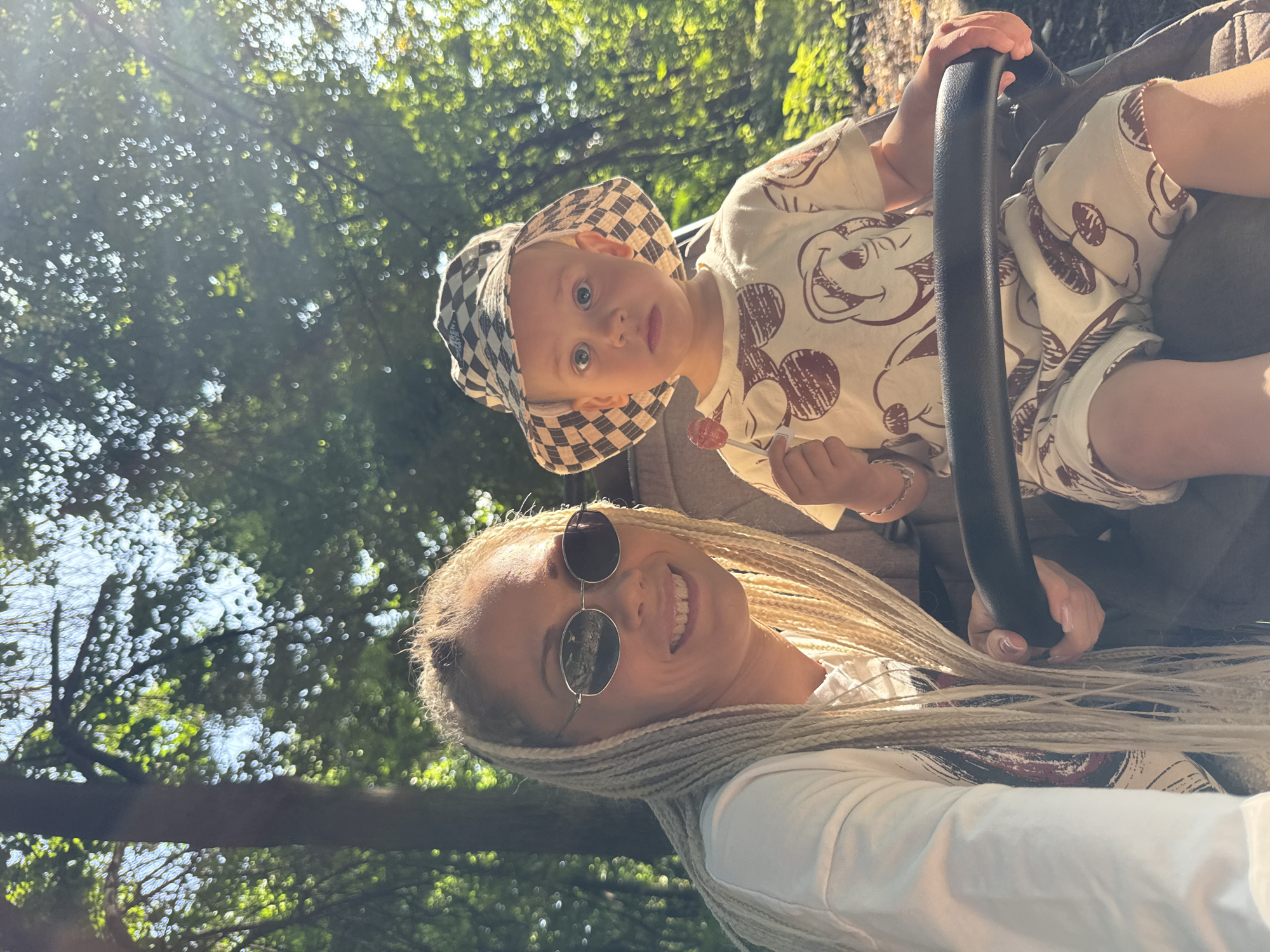 A woman with blonde braided hair and sunglasses is taking a selfie with a young child in a stroller. The child is wearing a checkered hat and a shirt with a Mickey Mouse print. The child is holding a lollipop. They are outdoors, with trees and sunlight in the background. The location is identified as Rusanovo, Russia.