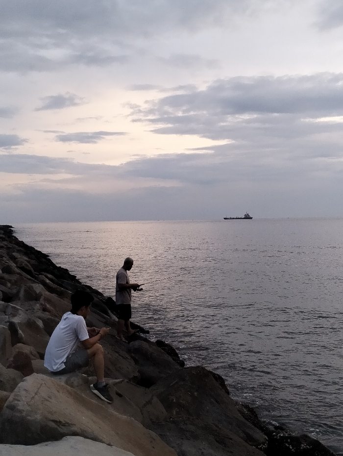 Two men are on a rocky breakwater along a coastline. One is seated, looking down at something in his hands. The other stands further out, holding a fishing rod. The water is a dark gray color and slightly choppy. In the distance, a large ship is visible on the horizon. The sky is overcast with gray and white clouds, hinting at either sunrise or sunset.