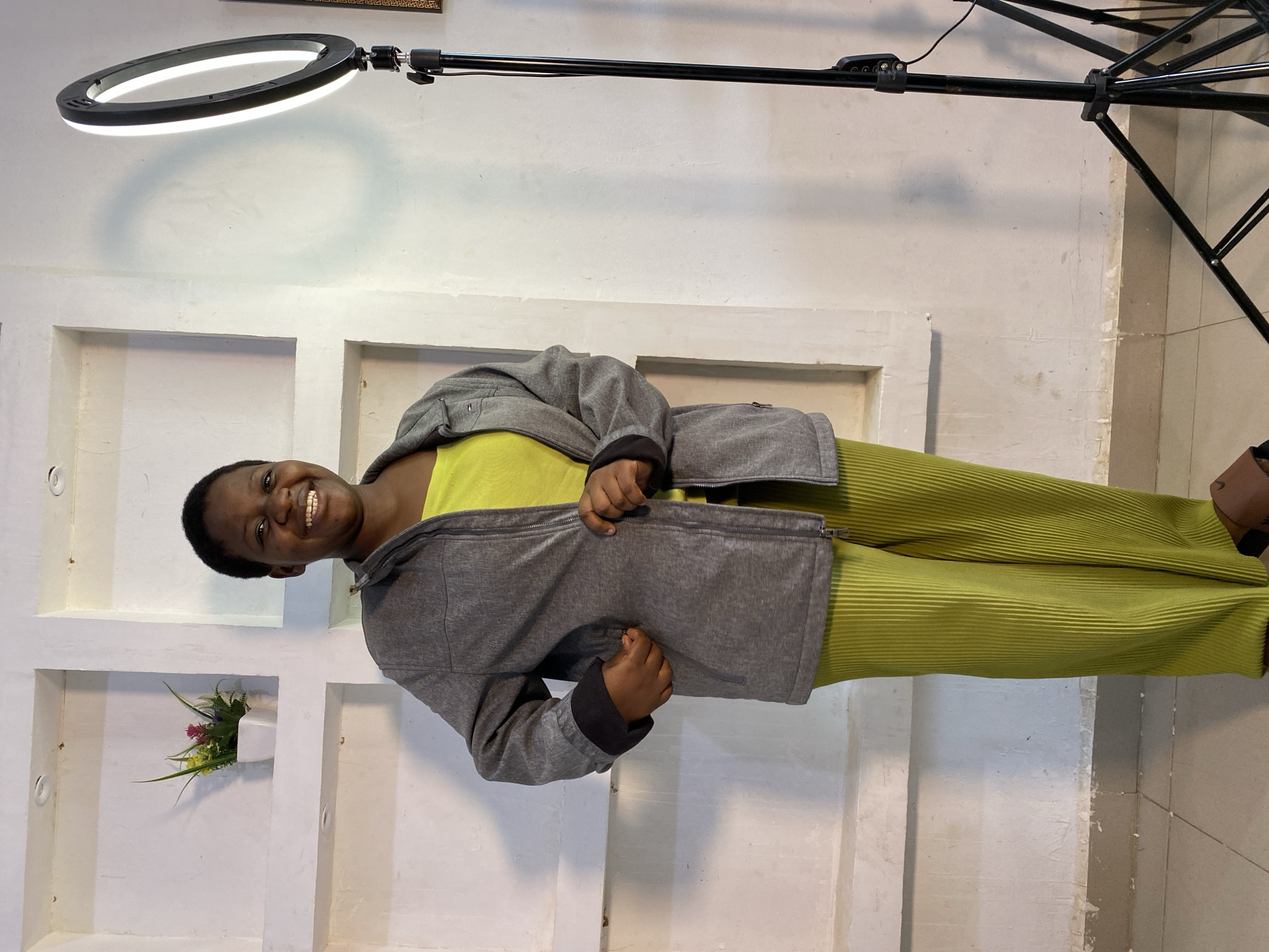 In an indoor setting, possibly in Jos, Nigeria, a young woman with dark skin and short, dark hair stands facing forward. She is smiling broadly and her head is tilted slightly to her right. She is wearing a bright yellow tank top under a grey zippered jacket. Her pants are a vibrant lime green and appear to be made of a pleated fabric. To her right, a tall, black ring light on a stand illuminates the scene. The background consists of a white wall with several recessed rectangular shelves. One shelf, to the woman's left, contains a small white pot with artificial green and pink flowers. The floor is tiled in a neutral color.