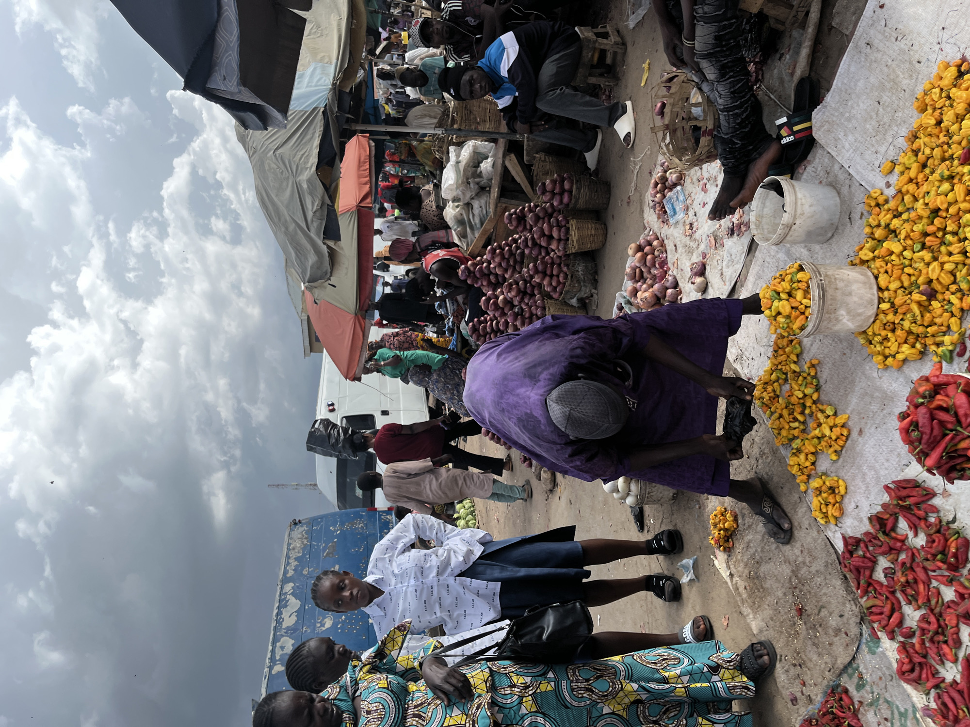 This is a street market scene in Rafin Par, Nigeria, under a cloudy sky. In the foreground, a person wearing a purple garment and a grey cap is bent over, sorting through yellow peppers. To the left, a pile of red peppers is spread out. Two people are standing in the mid-ground: one on the left wearing a colorful, patterned dress and carrying a black bag, and another to their right wearing a white shirt with black writing and a dark skirt. Behind them, several market stalls with awnings are visible. Various people are browsing and working in the market. In the background, a blue truck and a white van are parked. The ground is dirt and covered with some scattered produce and debris.