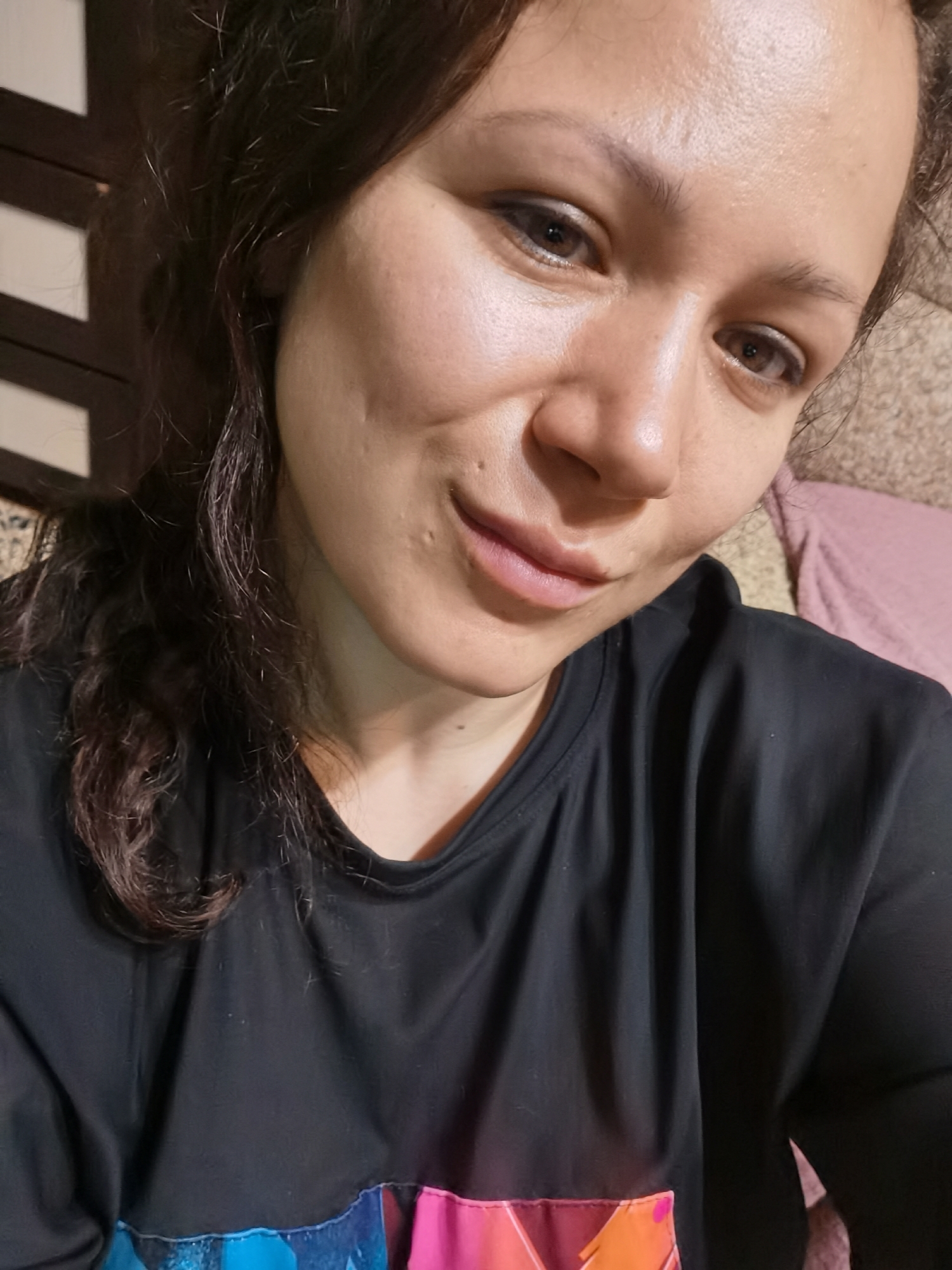 A close-up, selfie-style shot shows a woman from the chest up. She is looking directly at the camera with a slight smile. Her dark, wavy hair is pulled back from her face. She is wearing a black t-shirt with a colorful, abstract pattern on the lower portion of the shirt. The background is blurred and appears to be an indoor setting with a textured surface on the right and a dark, slatted structure on the left.