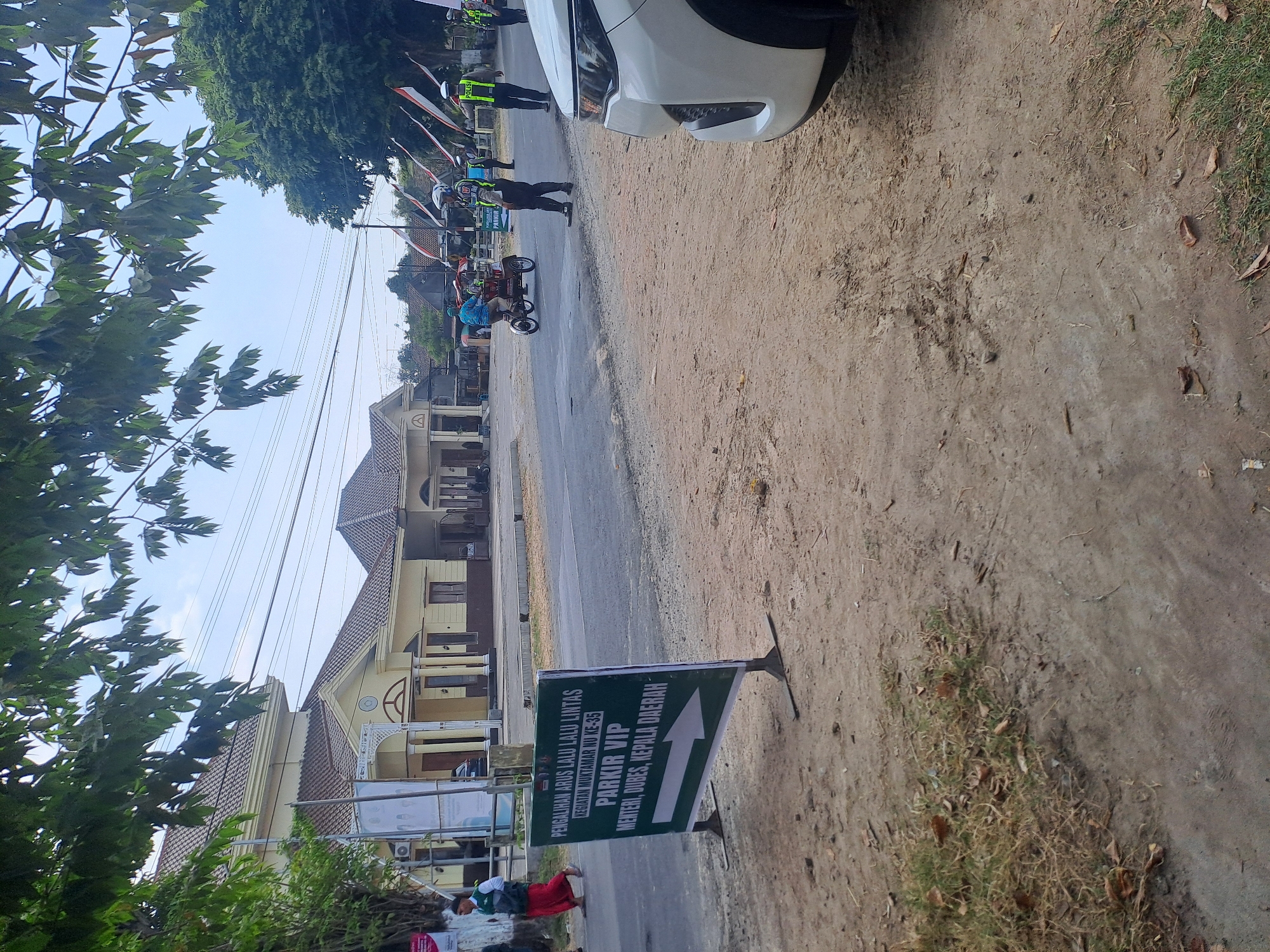 A green sign with white lettering is in the foreground, directing traffic for "PARKIR VIP MENTERI, DUBES, KEPALA DAERAH" and an arrow pointing to the right. In the background, a yellow building with a brown roof is visible, possibly a government building. Several police officers in uniform are present, with one directing traffic. A small, three-wheeled vehicle is being ridden by a person. Indonesian flags are displayed on poles in the background. The scene appears to be a road in Mojokrapak, Indonesia, with some trees and foliage visible. The ground is unpaved and dusty, with some patches of dry grass. A white car is partially visible on the right side of the image.