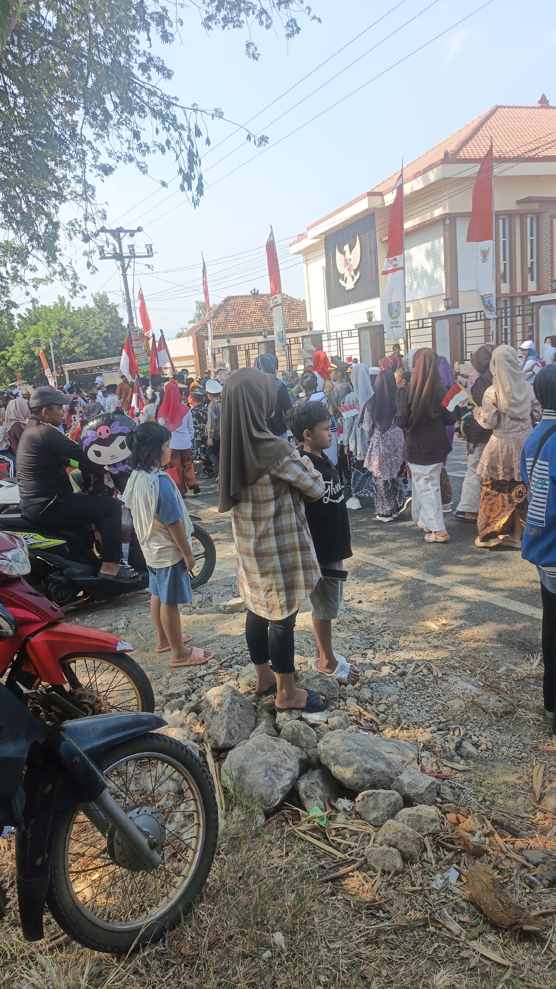 The image shows a street scene with a crowd of people, many of whom are children, gathered in front of a building. Red and white flags are visible, suggesting a patriotic event or celebration. Several motorcycles are parked on the side of the road. The building in the background appears to be a public institution, possibly a government office, with a crest or emblem displayed on its facade. The ground is a mix of asphalt and gravel, with some scattered rocks and dry vegetation. The lighting suggests it is daytime, with the sun casting shadows. The people are dressed in a variety of clothing, with some children wearing what appear to be costumes or uniforms. Some individuals are holding small flags. The overall atmosphere seems to be one of a community gathering or parade.