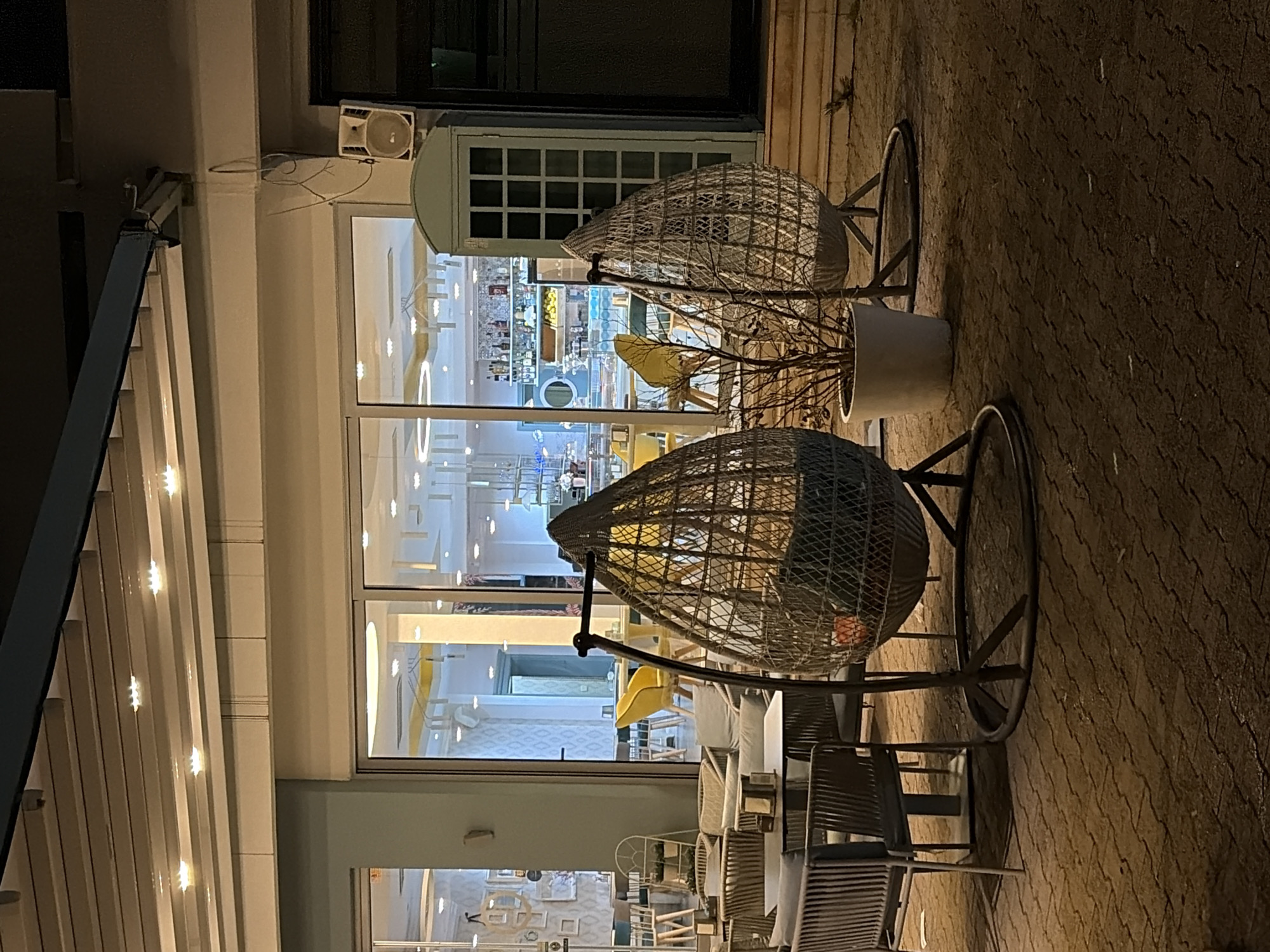 The image depicts an outdoor seating area of a restaurant or café, likely at night, in what appears to be Bar, Montenegro, based on the architecture and ambiance. The scene is dominated by two round, woven-type chairs, with their metal frames visible. In one of the chairs there is a potted plant. Behind the chairs, a large glass window reveals the interior of the establishment, showing a brightly lit area with various tables, chairs, and a bar with liquor bottles. The outdoor floor is paved with patterned concrete blocks, indicating a walkway or patio. To the left, a metal awning or canopy is partially visible, with built-in lighting. A speaker is mounted near the top corner of the window.
