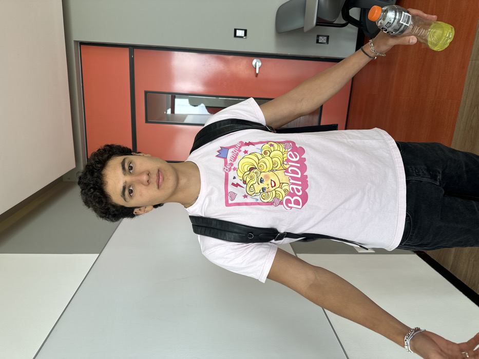 An eye-level photograph depicts a young adult male standing indoors against a two-toned wall and an orange door. The subject has dark, curly hair and is looking directly at the camera with a neutral expression. He is wearing a light pink short-sleeved t-shirt featuring a central graphic of the "Barbie" doll logo with blonde hair, accompanied by the text "Be unique." He wears dark-colored trousers and a black backpack over both shoulders. Multiple silver-colored bracelets and a white band are visible on his left wrist. His right wrist displays a dark-colored braided bracelet and a silver-colored chain bracelet. In his right hand, he holds a partially full, clear plastic bottle containing a yellow-green liquid, topped with a grey cap and an orange spout. The background includes a plain white wall on the left, transitioning to a grey section overhead. To the right, an orange door with a narrow vertical window is visible, reflecting a distorted image of a person. A grey wall panel is adjacent to the door. The floor is a solid orange color. A portion of a grey chair is visible in the lower right corner of the frame. This scene is located in Fraccionamiento Los Heroes Queretaro, Mexico.
