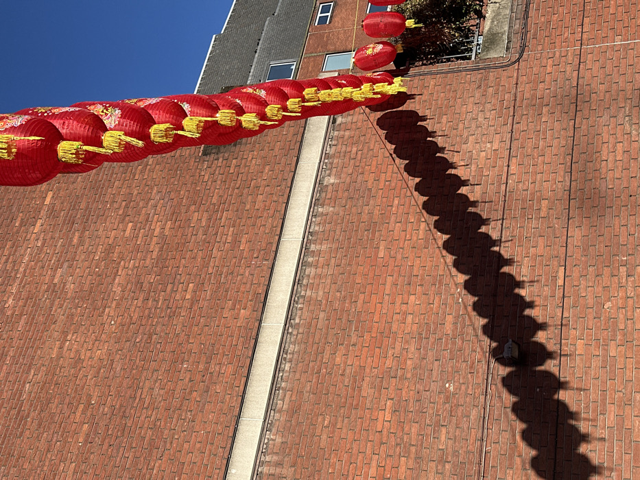 The image depicts a row of approximately twenty-six red Chinese lanterns with yellow tassels suspended horizontally across a sunny urban environment, identified as Chinatown, London, United Kingdom. The lanterns cast distinct, elongated shadows onto a textured red brick wall that dominates the left and bottom portions of the frame. A horizontal concrete band separates two sections of the brick wall. In the upper right quadrant, a section of a building with a grey sloped roof and several rectangular windows is visible against a clear blue sky. No individuals are present in the scene. The lighting suggests bright daylight conditions.