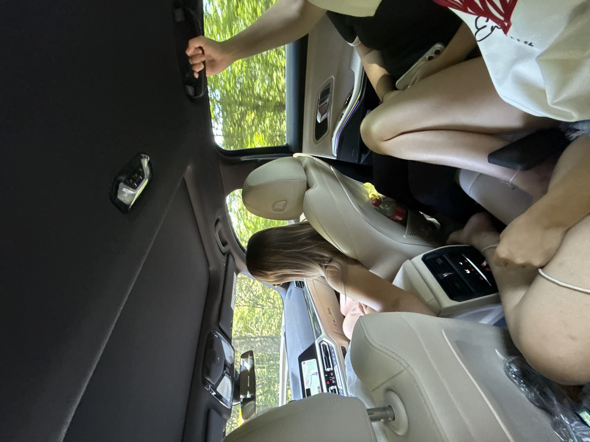 The image captures the interior of a car, likely a BMW based on the dashboard display and styling, as it travels through a wooded area. Three individuals are visible. A woman with blonde hair is in the driver's seat, looking towards the front. In the rear passenger seats, a person's arm is raised, holding onto a grab handle. Another person's legs are visible in the foreground, resting on the seat. The car's interior features beige leather seats and ambient lighting. Outside the windows, lush green trees indicate a rural or natural setting, consistent with the location of Oituz, Romania, known for its scenic beauty and proximity to forests. The car appears to be on a paved road, indicated by the navigation system displaying a map.