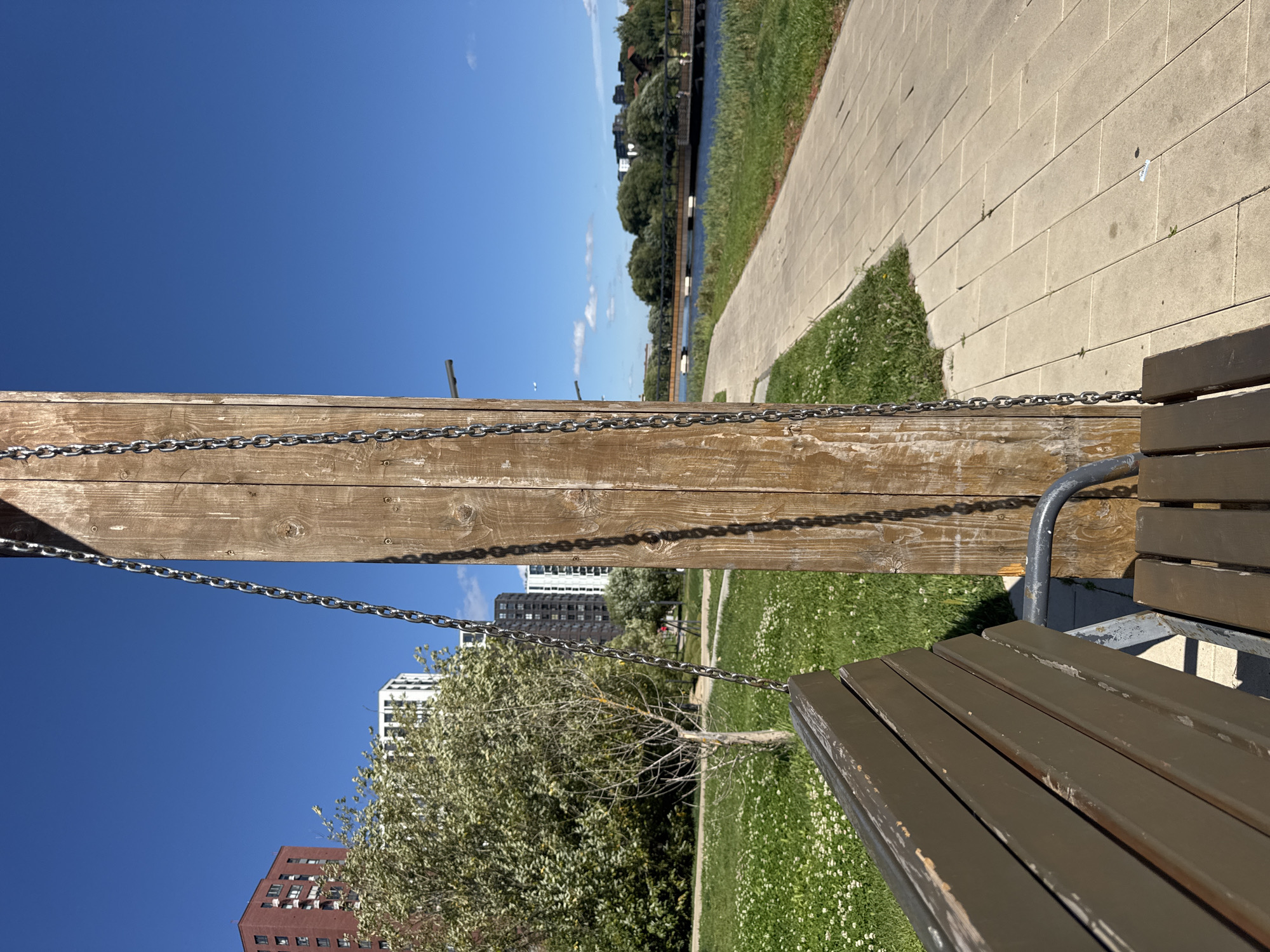 In the city of Kommunarka, Russia, a wooden swing set stands next to a paved walkway by a body of water. The swing, made of dark brown wooden slats, is suspended by metal chains attached to a thick wooden post. In the background, lush green grass dotted with small white flowers covers the ground. A paved walkway stretches alongside the water, with trees and residential buildings lining the opposite bank. The sky above is a clear blue. The image appears to be taken on a sunny day.
