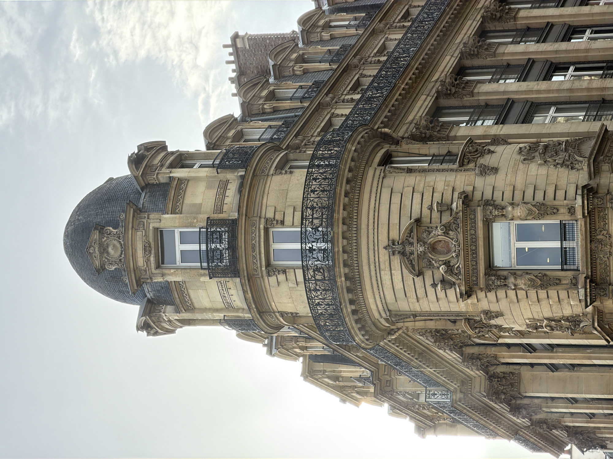 The image captures the ornate facade of a building in Paris, France. The building features a domed corner section with a slate roof and intricate stonework. The facade has multiple stories, with balconies adorned with wrought-iron railings. The architecture showcases classical elements, including window frames and decorative carvings. The sky is visible in the background.