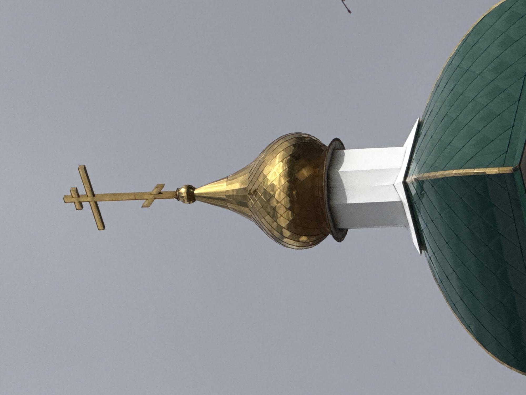 The image depicts the upper portion of a Russian Orthodox church, specifically the dome and cross. The dome is green and ridged, with a gold trim. A gold, onion-shaped spire sits atop a white, geometrically-shaped support, with a golden Orthodox cross at its apex. The background is a pale grey sky. This is likely located in the city of Chaikovsky, Russia.