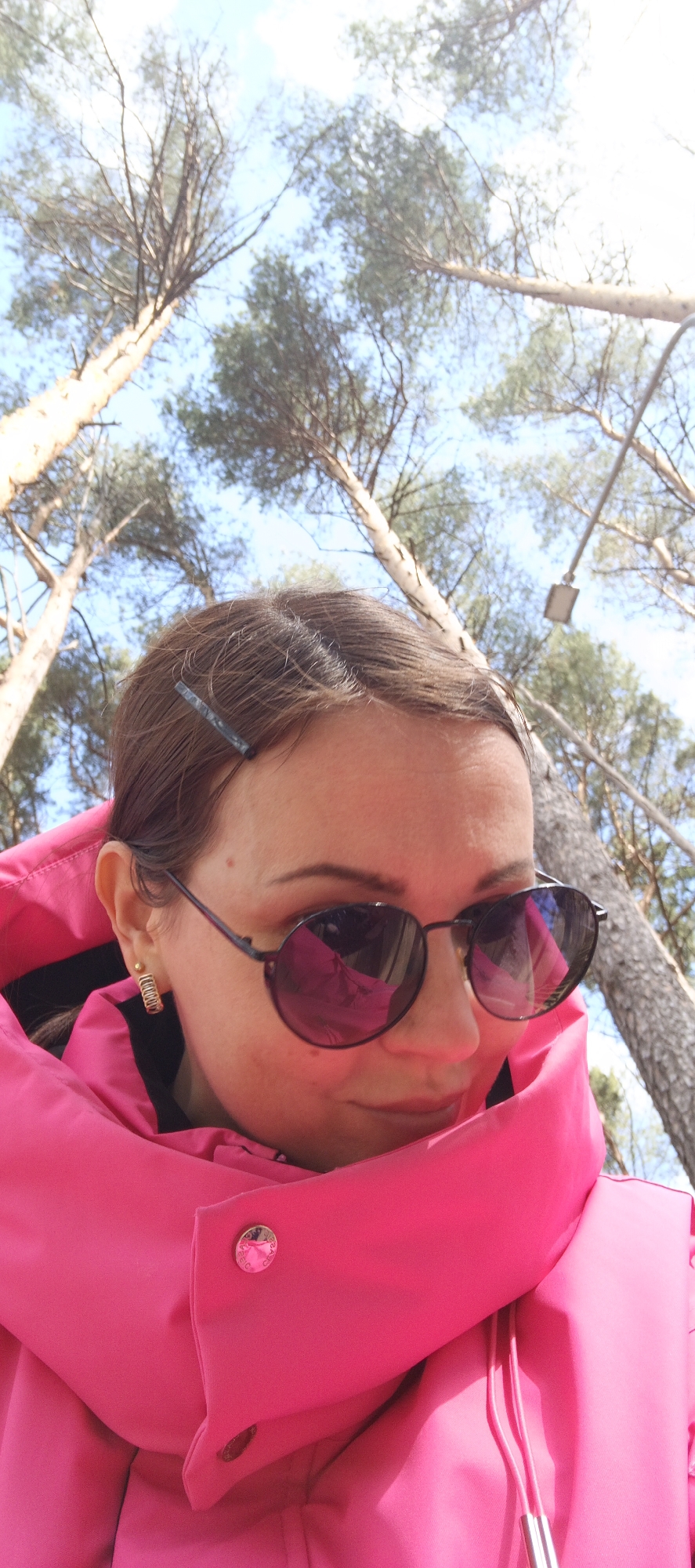A woman is pictured from a low-angle perspective, wearing sunglasses and a bright pink coat with a large collar drawn up around her face. Her hair is brown, styled with a hair clip. She has gold hoop earrings.
The background reveals a forest setting with tall trees stretching upwards, their branches sparse against a pale blue sky. A streetlight is visible.
The image was taken in Chaikovsky, Russia.