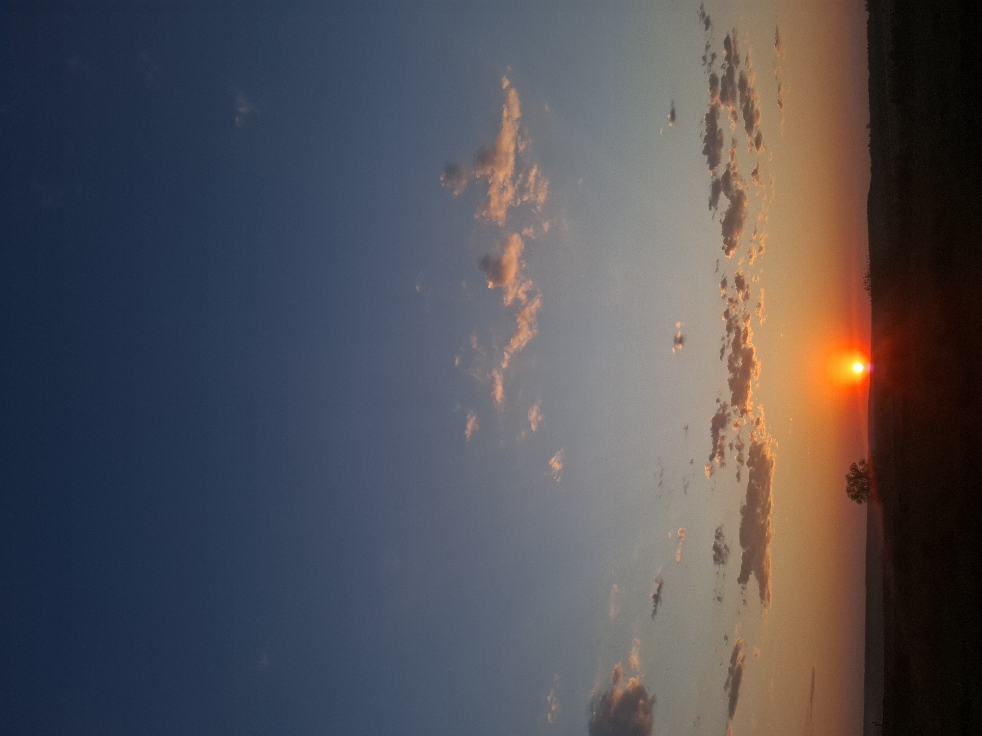 A sunset over a field in Khlevnoye, Russia. The sun is low on the horizon, casting an orange glow over the sky. Clouds are scattered across the sky, and a tree is visible in the foreground. The sky is a gradient of blue to orange, and the field is dark and indistinct.
