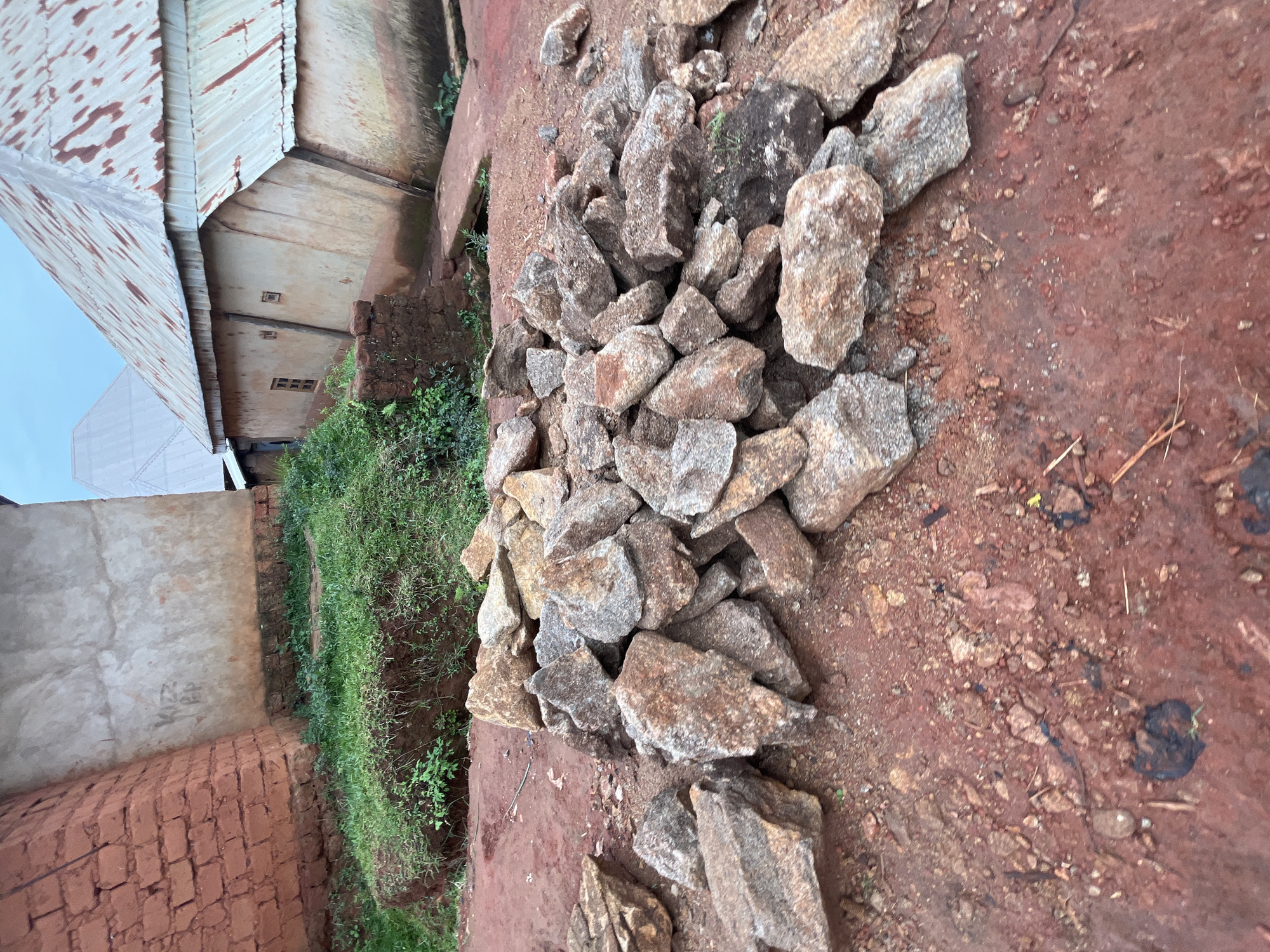 This media file depicts an outdoor scene in Gembu, Nigeria. The primary subjects are a pile of rough, irregularly shaped rocks of varying sizes and colors, predominantly gray and brown with some reddish hues. These rocks are scattered across a reddish-brown, earthy ground.
To the left of the rocks, there is a patch of vibrant green grass and some small plants growing along a slight incline. Further to the left, a building is partially visible. It appears to be constructed with brick at the bottom, transitioning to a concrete wall above. A corrugated metal roof, showing signs of rust, extends from this building. The overall impression of the setting is one of a rural or semi-developed area, possibly a construction site or a place where materials are gathered.
There is no discernible activity or event taking place in the image; the scene is static, suggesting a moment captured in time. The lighting indicates it is daytime, though the sky is overcast and muted, suggesting a cloudy or perhaps early morning/late afternoon light. There are no people visible in the frame. No text is visible in the image. The overall mood is utilitarian and somewhat unkempt, common in areas where construction or natural materials are abundant.