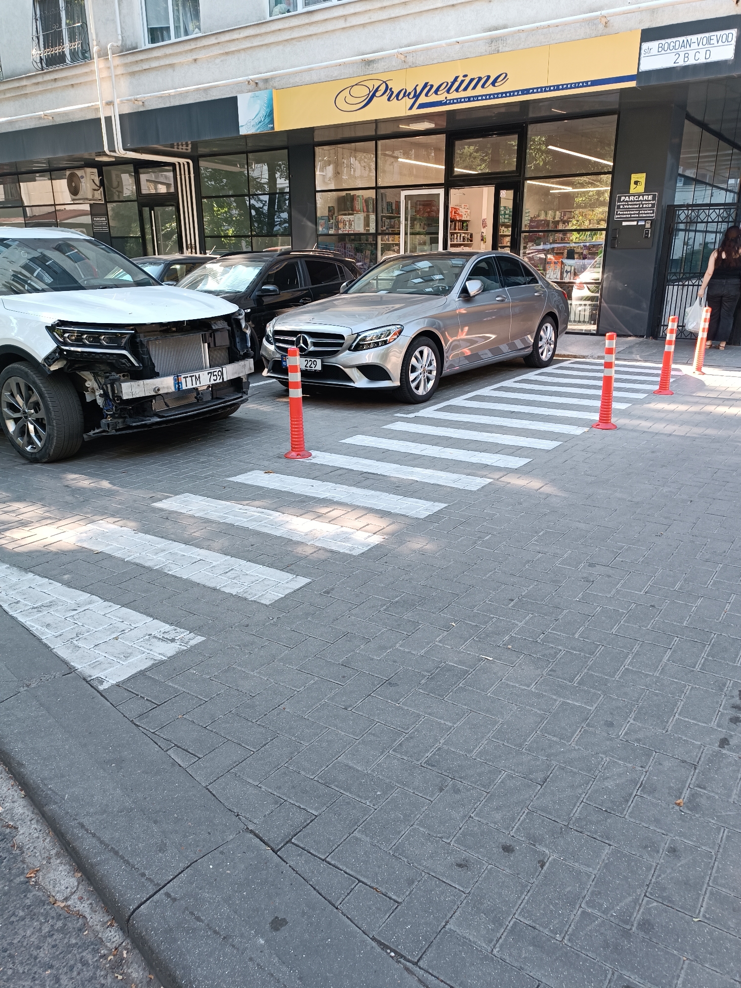 The image shows a street scene in Chișinău, Moldova, with two cars parked on a pedestrian crossing in front of a commercial establishment named "Prospetime". A white SUV with a damaged front end is on the left, and a silver Mercedes-Benz sedan is on the right. Several orange traffic cones are placed on the pedestrian crossing. The building facade features large glass windows, a prominent yellow sign with the establishment's name, and architectural details. A street sign indicates "str. BOGDAN-VOIEVOD 2 B C D". A woman with dark hair is visible walking away from the scene on the right. The ground is paved with grey interlocking bricks.
