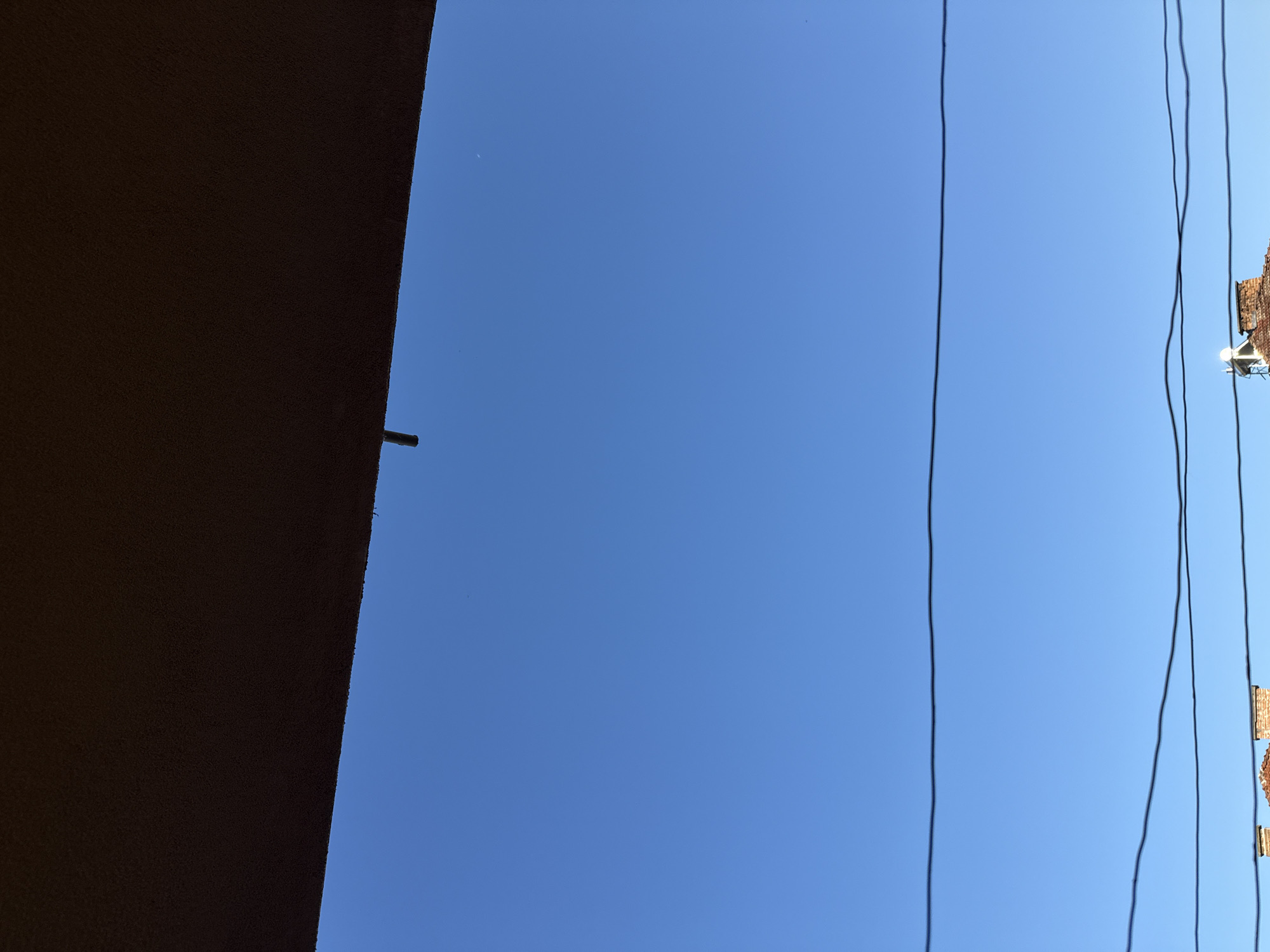 The image displays a clear blue sky with a portion of a building's overhang in the upper part. Below, several horizontal power lines stretch across the frame. At the bottom, the rooftops of buildings are visible, one featuring a chimney. The scene is set in the city of Draginovo, Bulgaria.