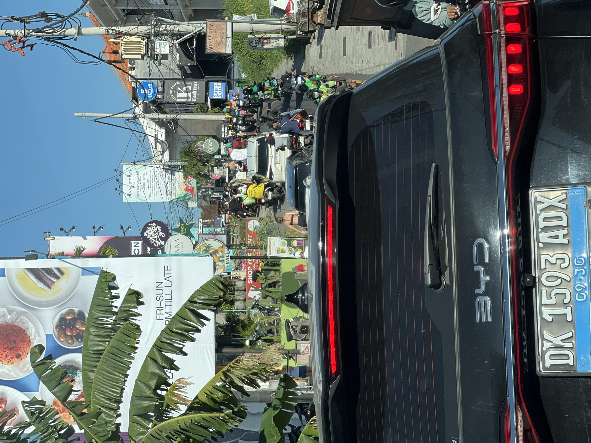 The image depicts a busy street scene in Bali, Indonesia, viewed from behind a black BYD car with license plate DK 1593 ADX. A traffic jam is visible, consisting of numerous motorcycles, cars, and other vehicles. Buildings line the street, adorned with a variety of signs and advertisements, including a large billboard promoting a restaurant with the text "FRI-SUN RM TILL LATE." The billboard also displays an advertisement for "DAY-ZERO GWK CULTURAL PARK APRIL 2026 BALI," and an ad for "Acai Shop Inaya." Lush tropical plants frame the lower portion of the image. Overhead are numerous electrical wires and utility poles.