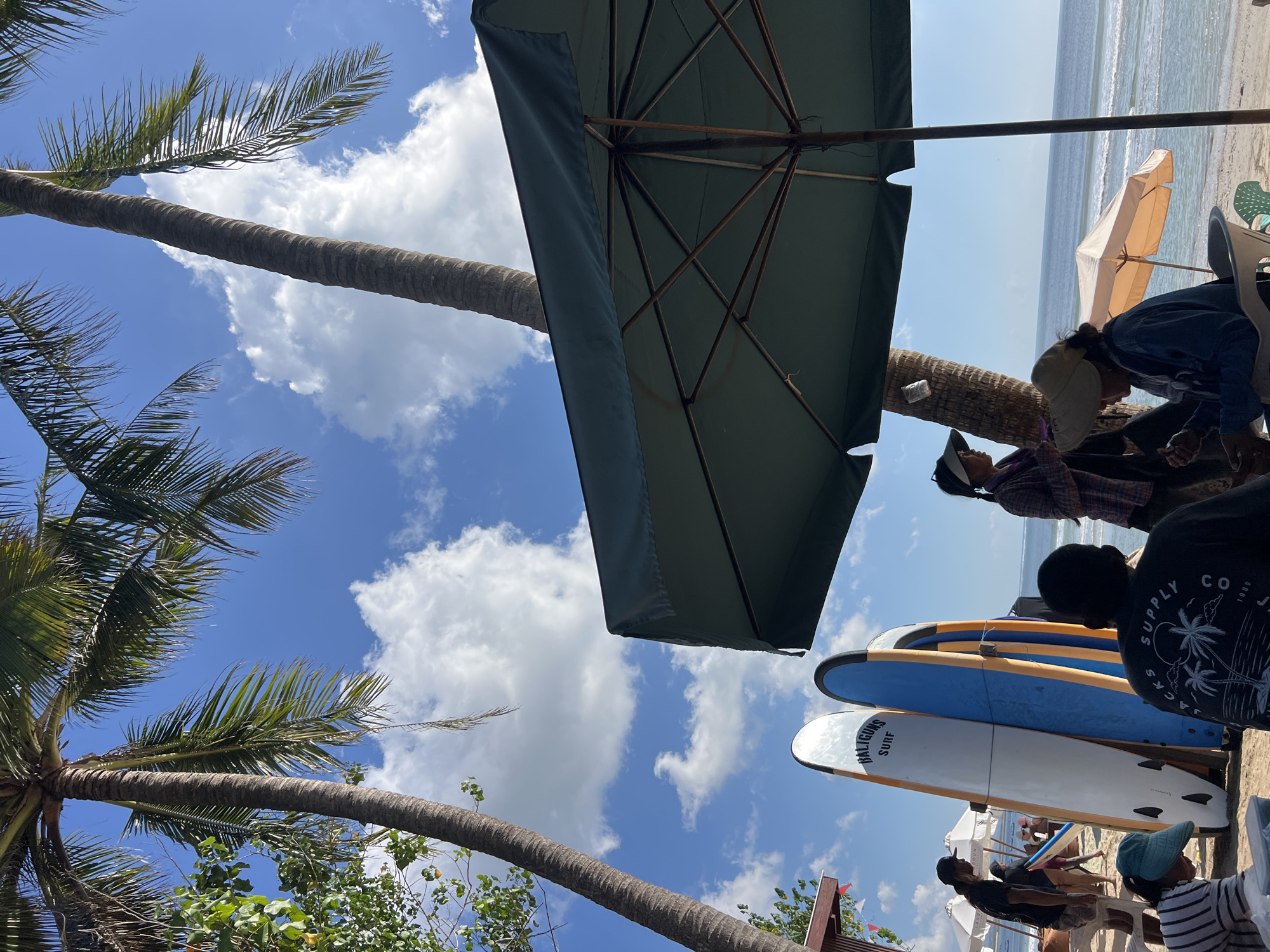 An outdoor, ground-level, eye-level shot on a sunny day at Kuta Beach, Indonesia, captures a group of people gathered near surfboards under palm trees. The image features a dark green beach umbrella in the foreground, obscuring a portion of the scene. Two surfboards, one white with "BALIGUNS SURF" printed on it and a blue one, stand upright. Several individuals are present: a man in a black t-shirt and shorts, another man in a black t-shirt with a palm tree graphic, a woman in a plaid shirt and a hat, and a woman in a beige hat and denim jacket. Some are interacting, with one woman appearing to offer something to another. The background shows the ocean with gentle waves, a clear blue sky with scattered white clouds, and more palm trees. A small orange umbrella is visible further down the beach. The overall atmosphere suggests a relaxed beachside setting.