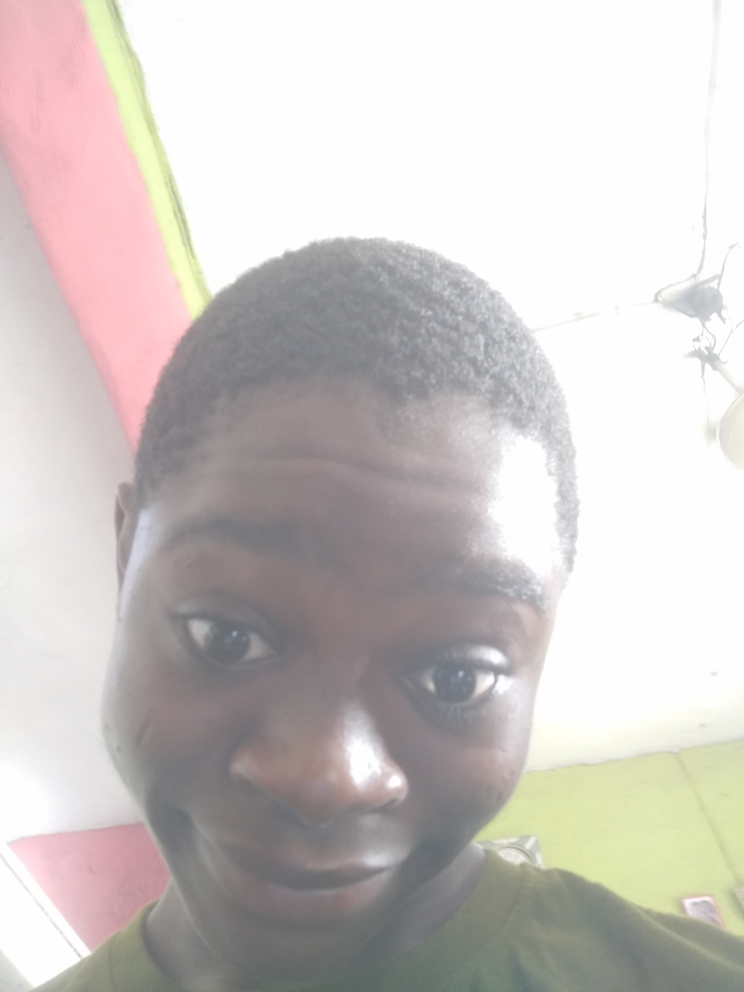 A close-up, slightly overexposed selfie photograph shows a person with dark skin, short dark hair, and a green shirt. The person is indoors, facing the camera with a slight smile. The background includes a ceiling with pink and green sections. An object resembling a light fixture is visible in the top right corner. The location is Shangisha, Nigeria.