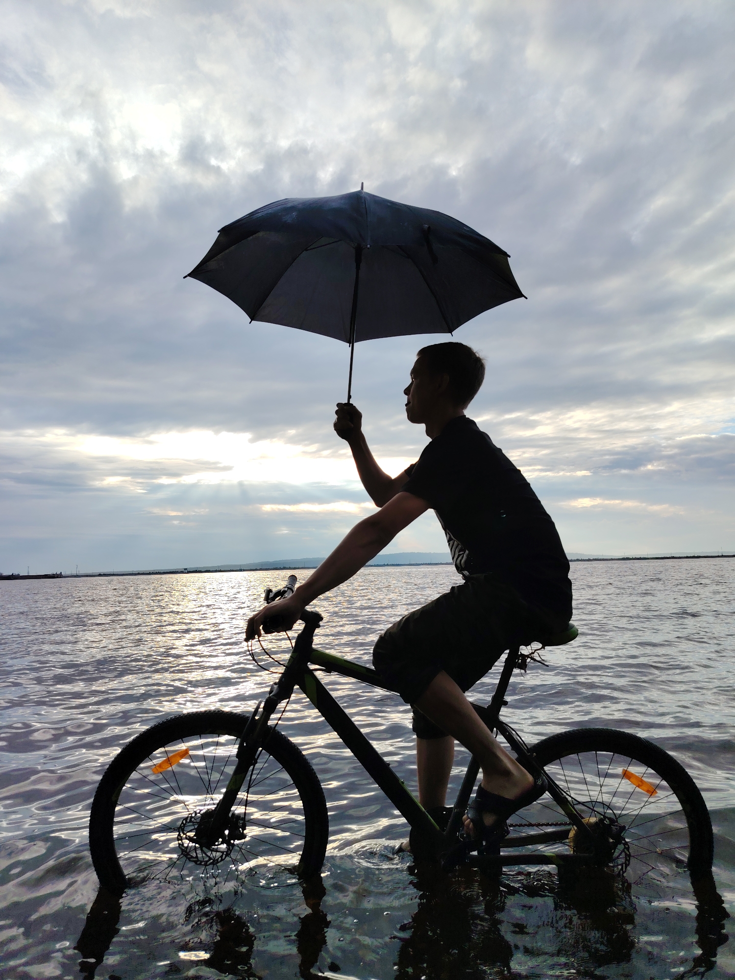 A young man, silhouetted against a cloudy sky, sits astride a bicycle in the shallow water of a body of water. He is holding a black umbrella over his head, shielding himself from the elements. The water reaches the tires of the bicycle, and the setting sun reflects on the rippling surface. The sky is overcast with dramatic clouds, suggesting either the end of a storm or the approach of one. The man is wearing a black t-shirt and dark shorts, and sandals on his feet. The scene evokes a sense of peaceful solitude and resilience. The location appears to be a large lake or sea near Chaikovsky, Russia, given the open water and distant shore. The time of day is likely late afternoon or early evening, as indicated by the subdued sunlight.