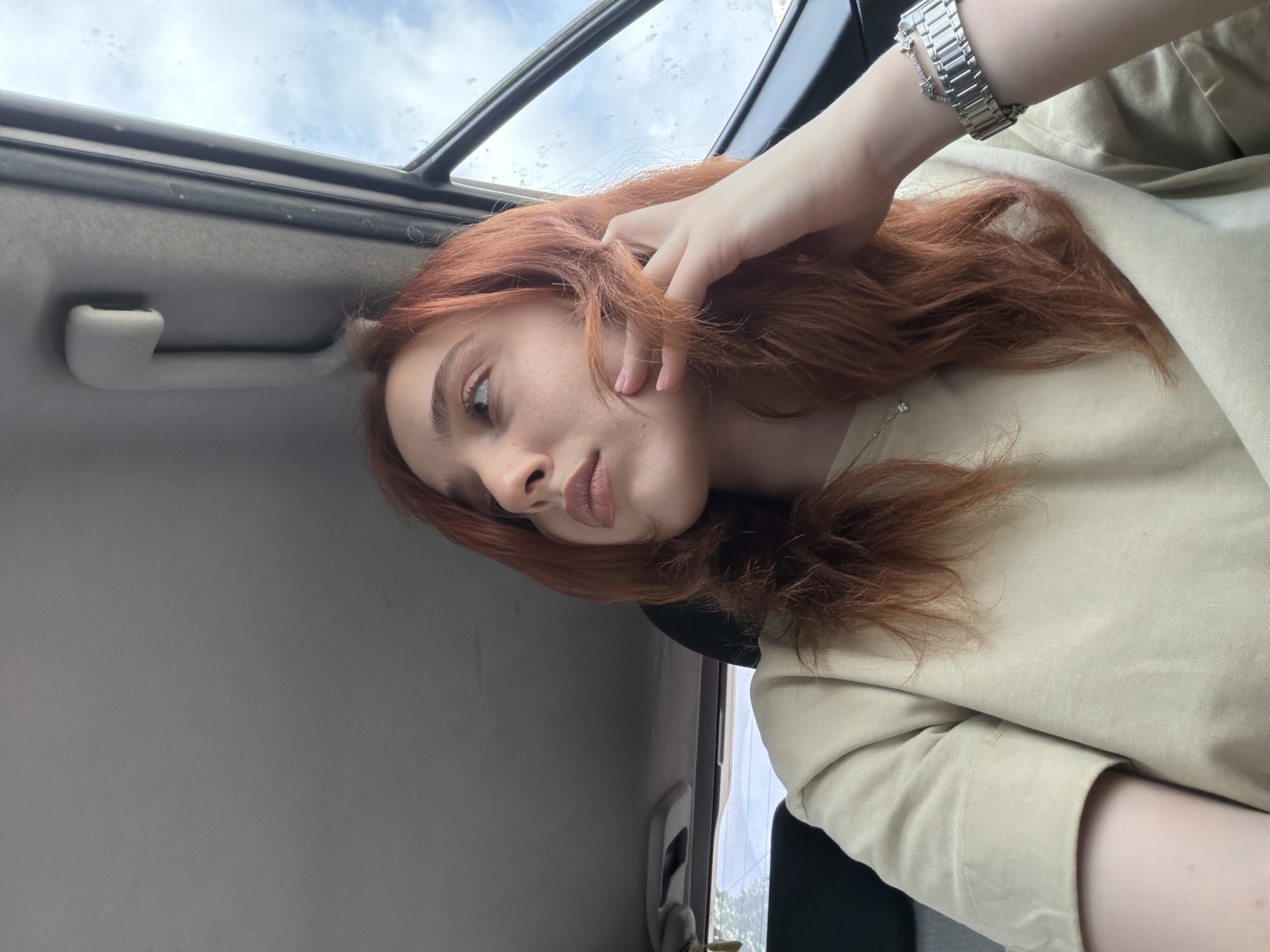 The image shows a fair-skinned person with red hair inside a vehicle. The person is wearing a light-colored shirt and a silver watch. They appear to be touching their hair with their hand. The interior of the vehicle includes a grey ceiling with a sunroof. The location is unknown and cannot be determined from the image, nor can it be determined that the location is Tbilisi, Georgia.
