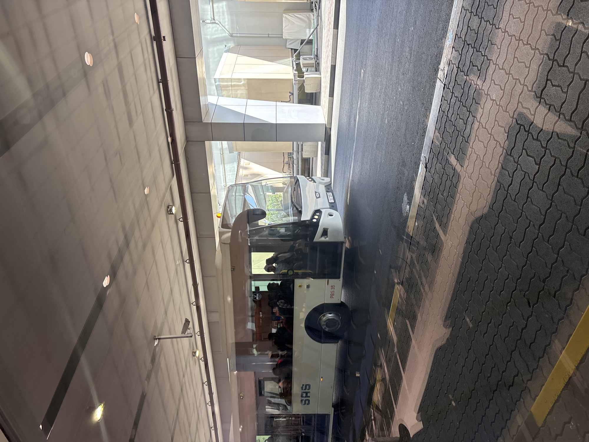 This image depicts a SAS Transport passenger bus in a sheltered outdoor transportation area, likely within Sharjah International Airport in Sharjah, United Arab Emirates. The scene is observed through a reflective surface, evident by distinct reflections of interior lights and structures on the upper portion of the image.
The central subject is a modern, white bus featuring "SAS" in blue lettering on its side and "PBS 35" near the rear wheel well. The front passenger door is open, revealing steps. Numerous individuals are visible inside the bus through its tinted windows and at the open doorway, suggesting passenger boarding or disembarking. Their attire appears casual.
The bus is parked on an asphalt road marked with white lines. To the right of the bus, a sidewalk is paved with dark grey interlocking pavers in a zigzag pattern, bordered by a yellow-painted curb. Strong, well-defined shadows, indicative of bright sunlight, are cast across the road and sidewalk from the overhead structure and potentially the bus itself.
Above, a flat concrete ceiling extends overhead, featuring recessed circular light fixtures, some illuminated. A dark-colored pipe runs horizontally along the ceiling, from which several hooks or attachments are suspended. Two CCTV cameras are mounted from the ceiling, angled downwards.
In the background, beyond the bus and behind large support columns, are multiple large, rectangular, light beige structures, likely industrial HVAC units or utility enclosures. Associated piping and smaller outdoor air conditioning units are visible around these structures.
