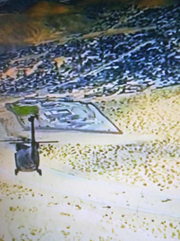 A helicopter flies over a desert landscape with an irregular pattern of light brown and beige textures. In the distance, a denser, blueish patch suggesting a settlement or urban area appears. A large, angular structure, potentially a military complex or base, is prominently situated within a lighter-colored, sand-like zone, contrasting with the surrounding ground. The entire scene has a blurry and indistinct quality.