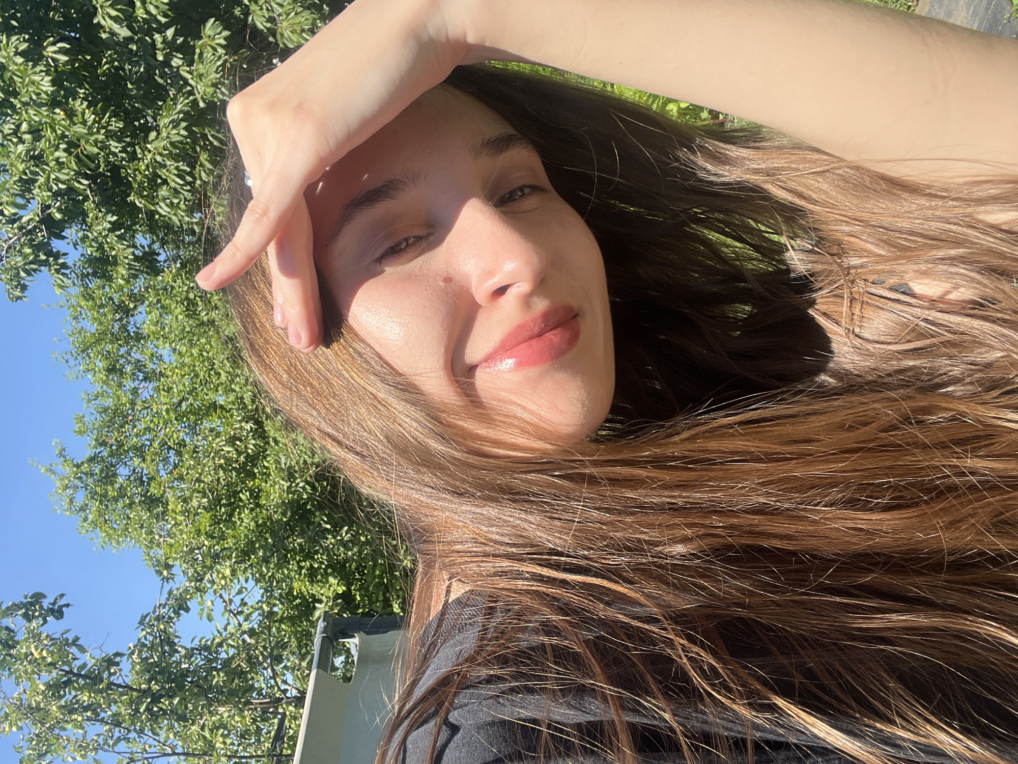 This media file is a close-up, outdoor selfie of a young person with long, light brown hair. The subject is looking towards the camera with their right hand raised to their forehead, partially obscuring their brow. Their eyes are mostly closed, and they have a slight smile on their face, with glossy, pink-toned lips. Sunlight is casting a strong shadow across their face, highlighting their cheekbones and the bridge of their nose. Their skin appears to have a dewy glow.
The background is a blur of green foliage, suggesting a natural, outdoor setting. The sky above is a clear, bright blue. A portion of a dark garment is visible at the bottom of the frame. The overall impression is one of warmth and relaxation, perhaps taken on a sunny day. The time of day appears to be daytime, given the bright sunlight and clear sky. There is no visible text in the image.
